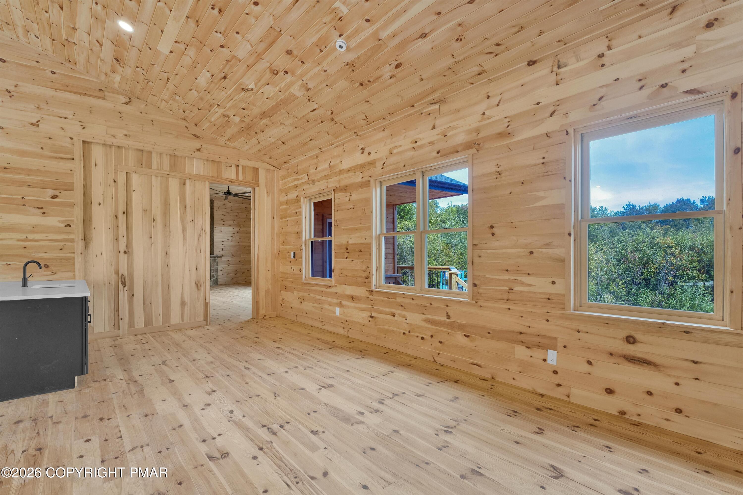Unfurnished knotty pine room with a vaulted ceiling, wood floors, and windows overlooking trees.