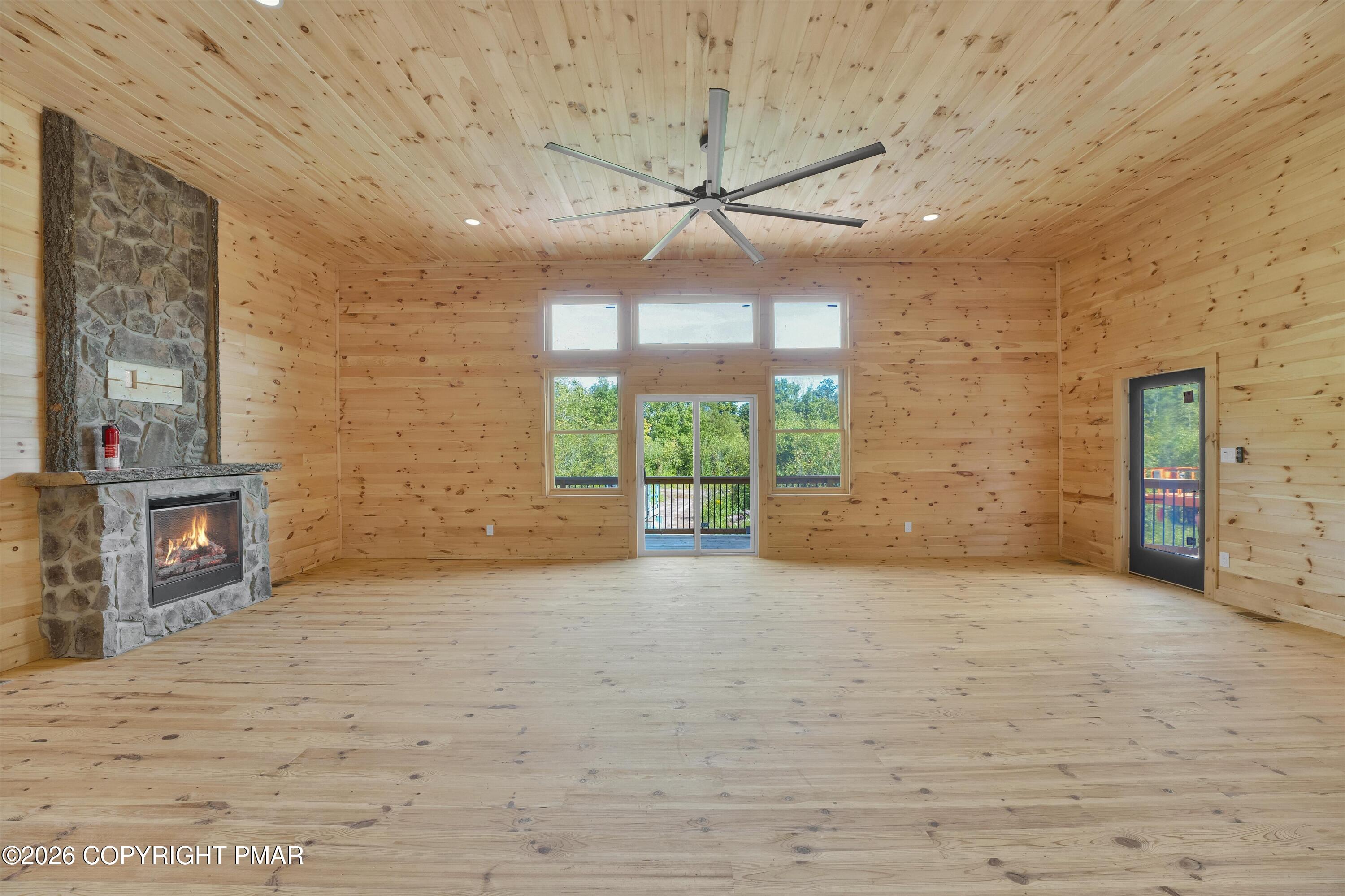 Empty pine-paneled living room with a stone fireplace, large ceiling fan, and windows overlooking a deck.