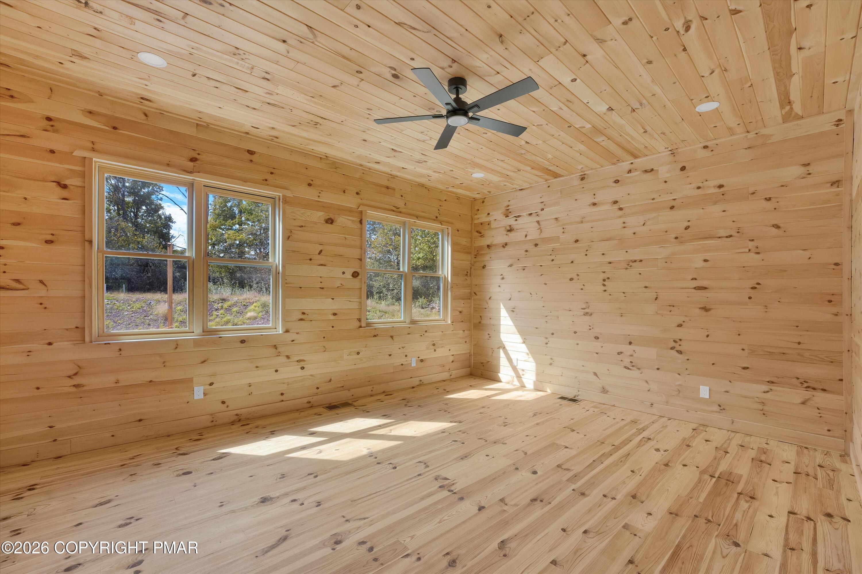 Empty knotty pine room with two sets of windows and a dark ceiling fan.