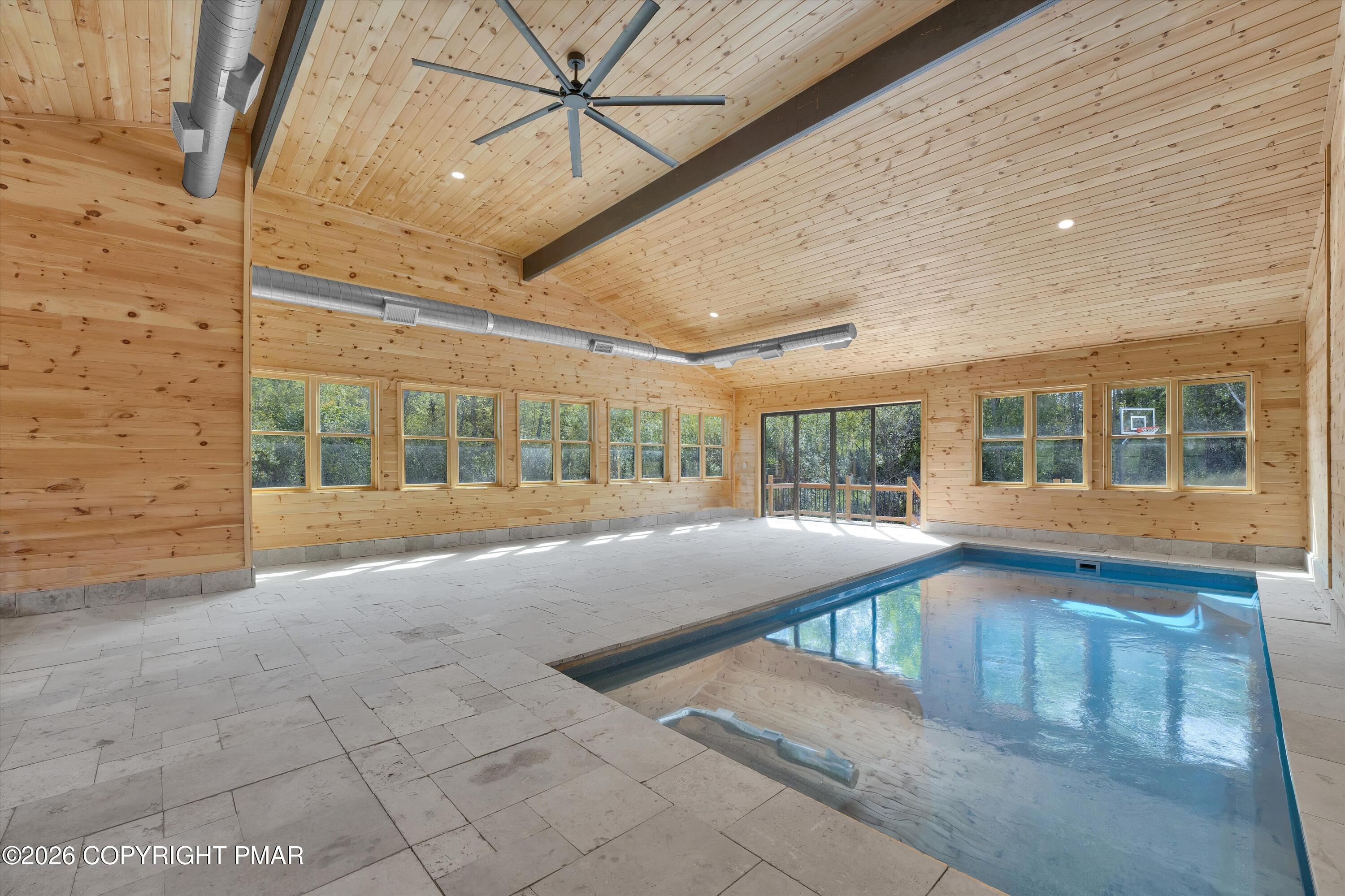 Indoor pool with a wood-paneled ceiling, large windows, and pale stone-tile deck.