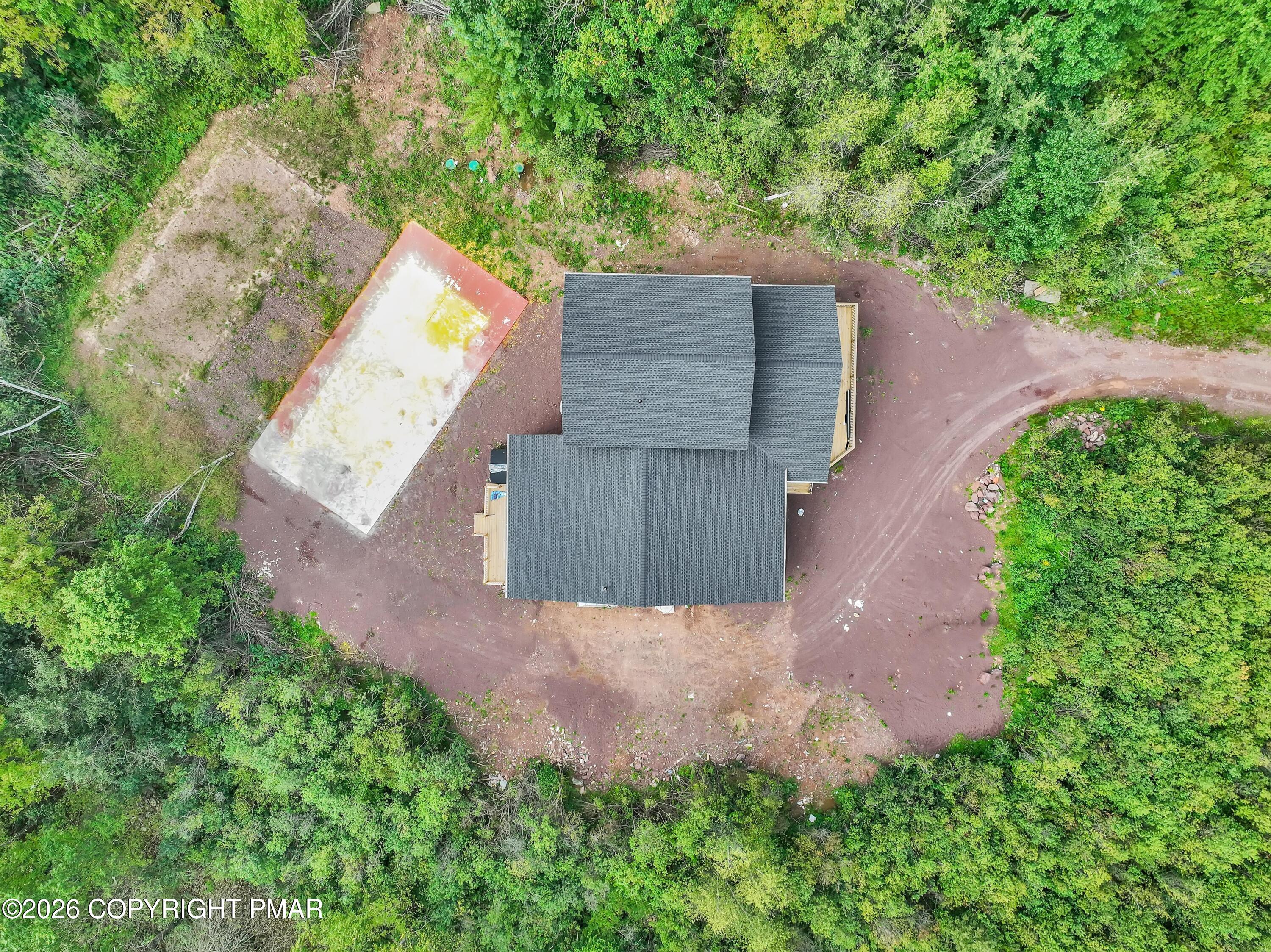 Aerial view of a house in a wooded clearing, with a curving dirt driveway and large paved area.