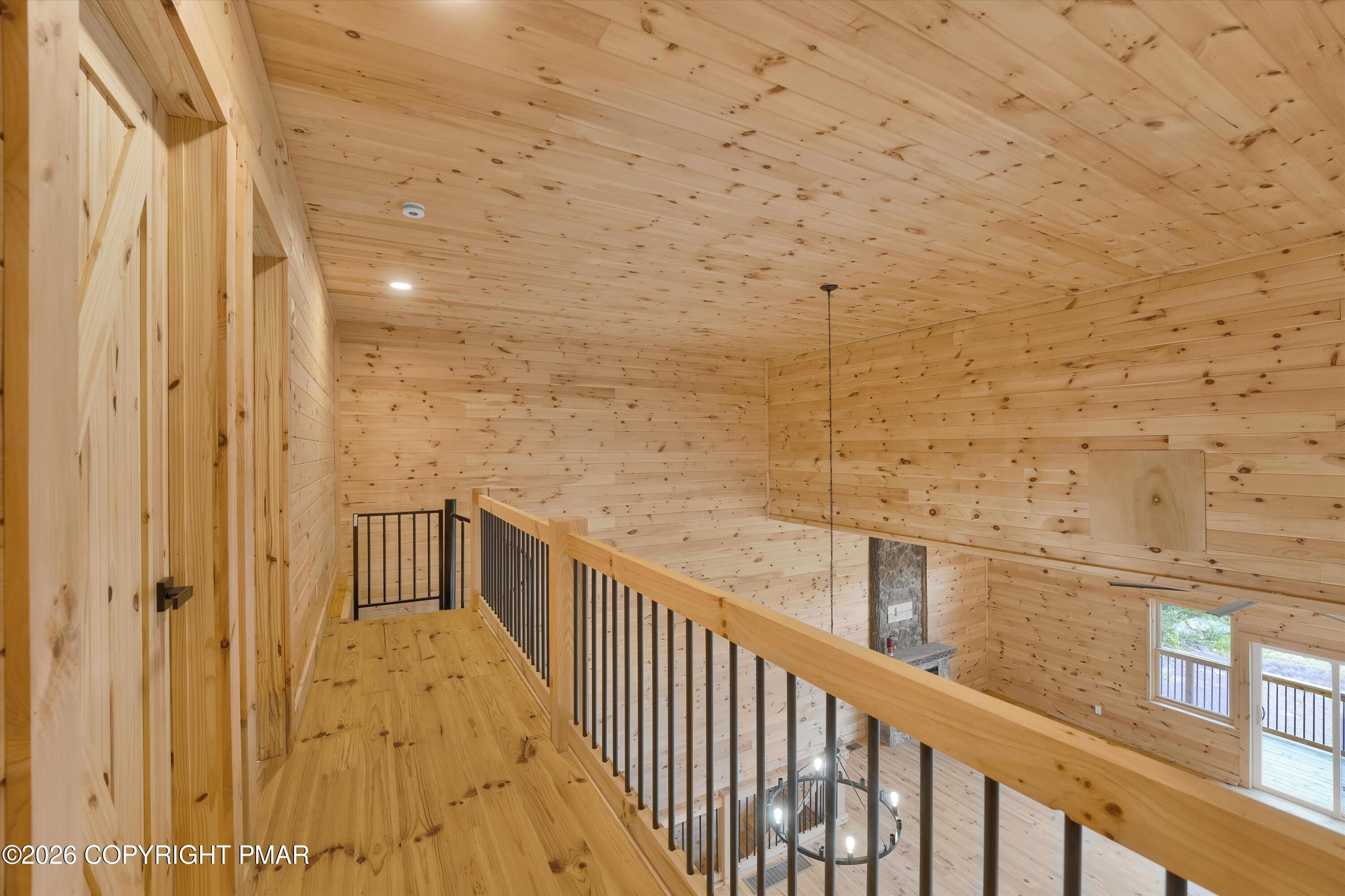 Wood-paneled loft hallway overlooking a living room, with a black metal railing and stone fireplace.
