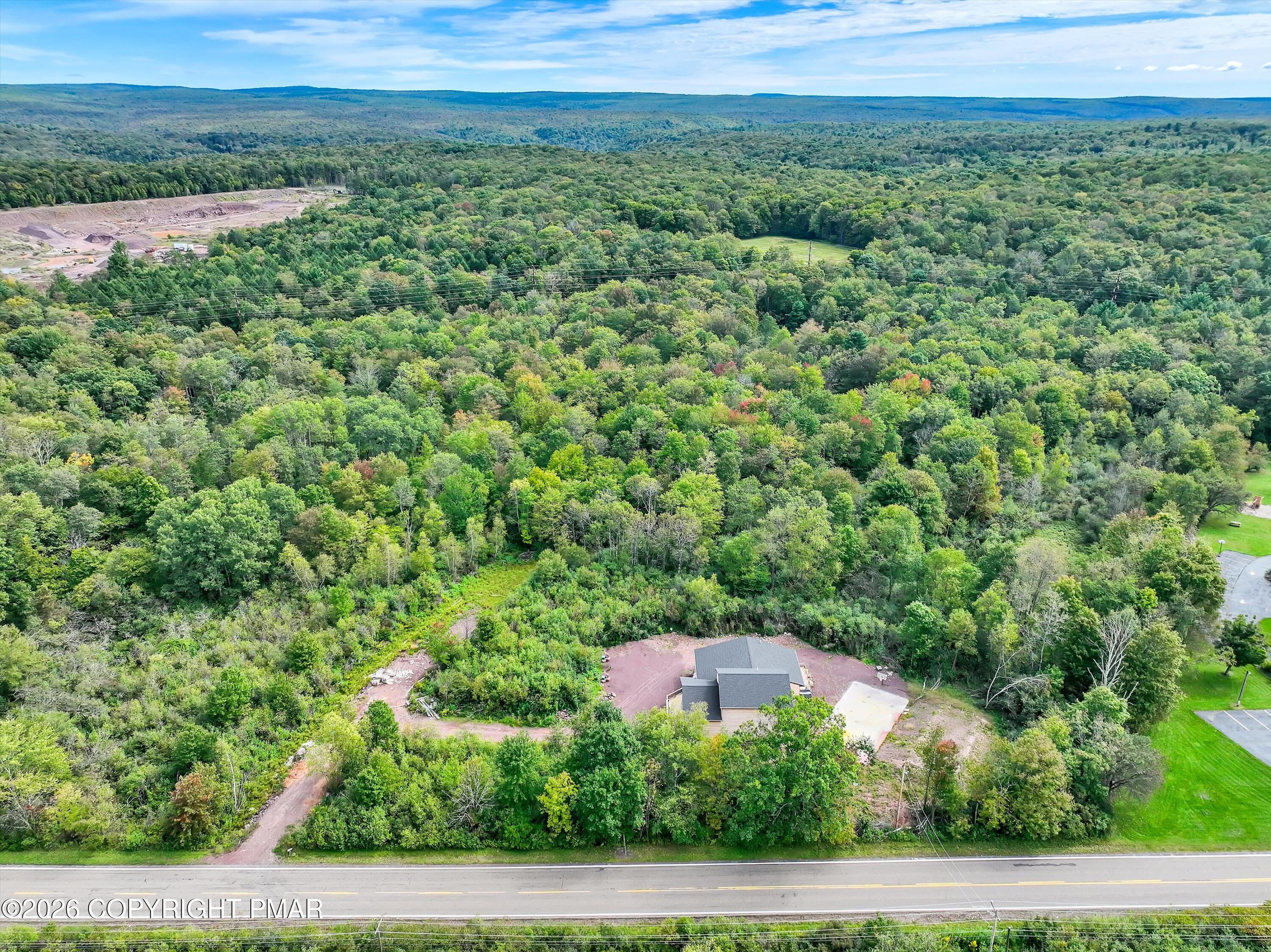 Aerial view of a house amid dense woods, with a dirt driveway and forested hills beyond.