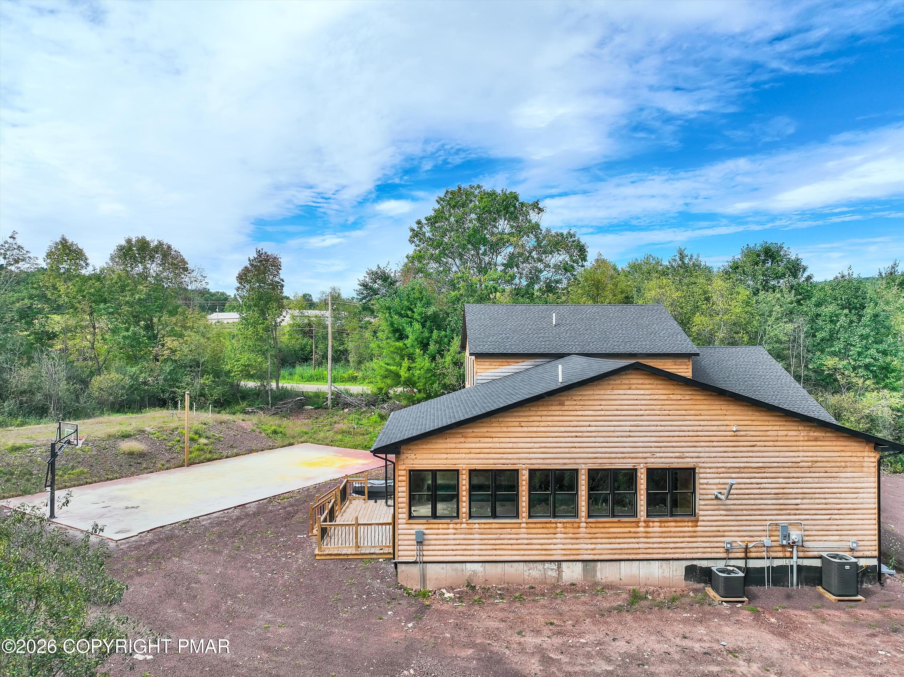 Rear of a log-sided house with a deck and adjacent basketball court, surrounded by trees.
