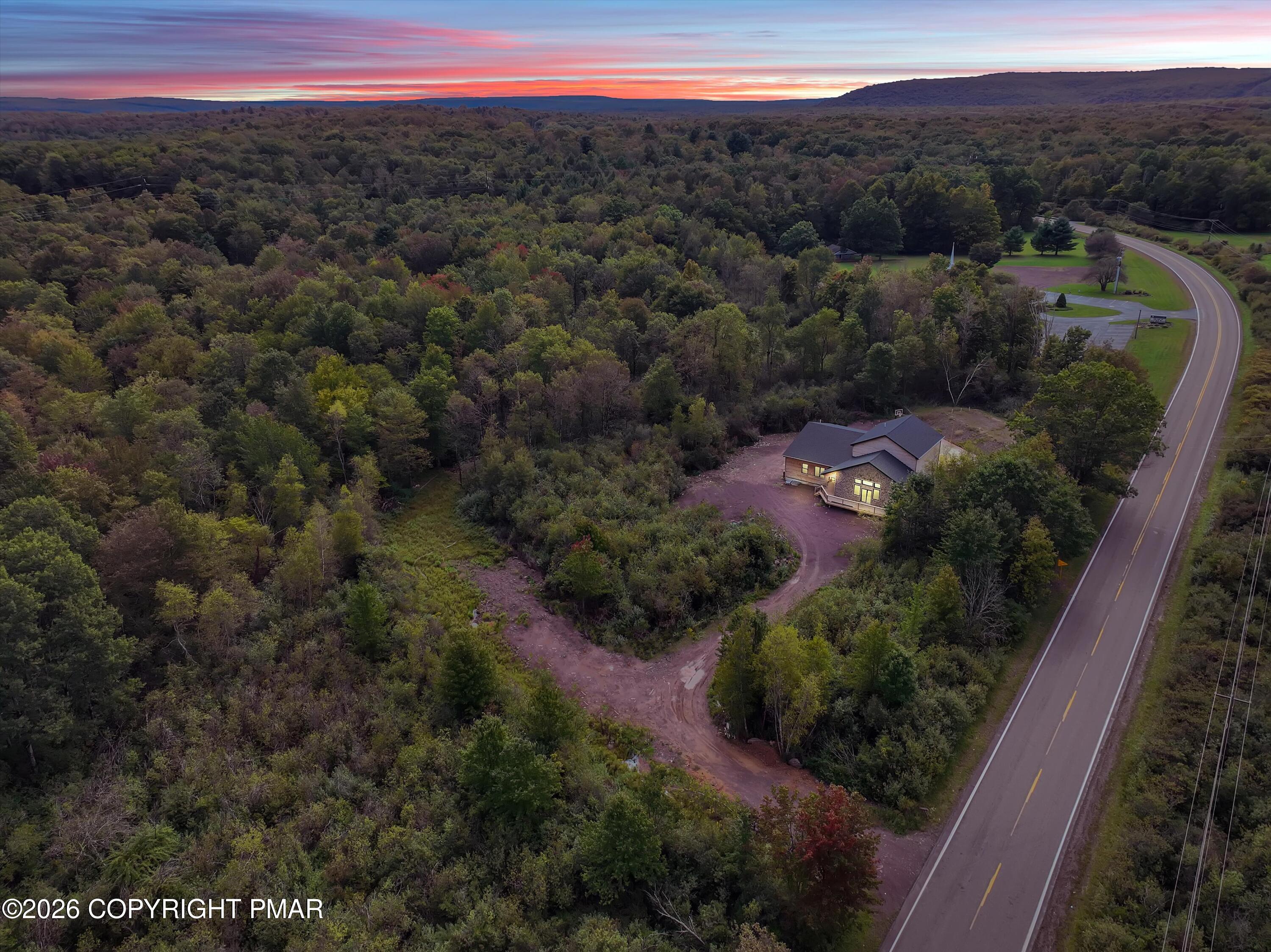 Aerial view of a house among dense autumn woodland, beside a curving road at sunset.