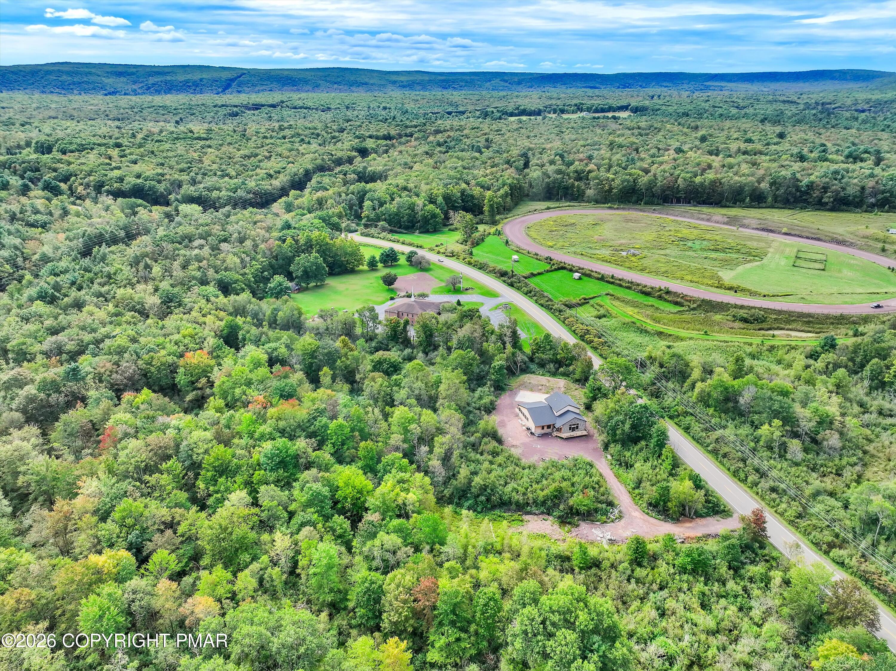 Aerial view of a partially built home amid woodland, with a curving road and oval track nearby.