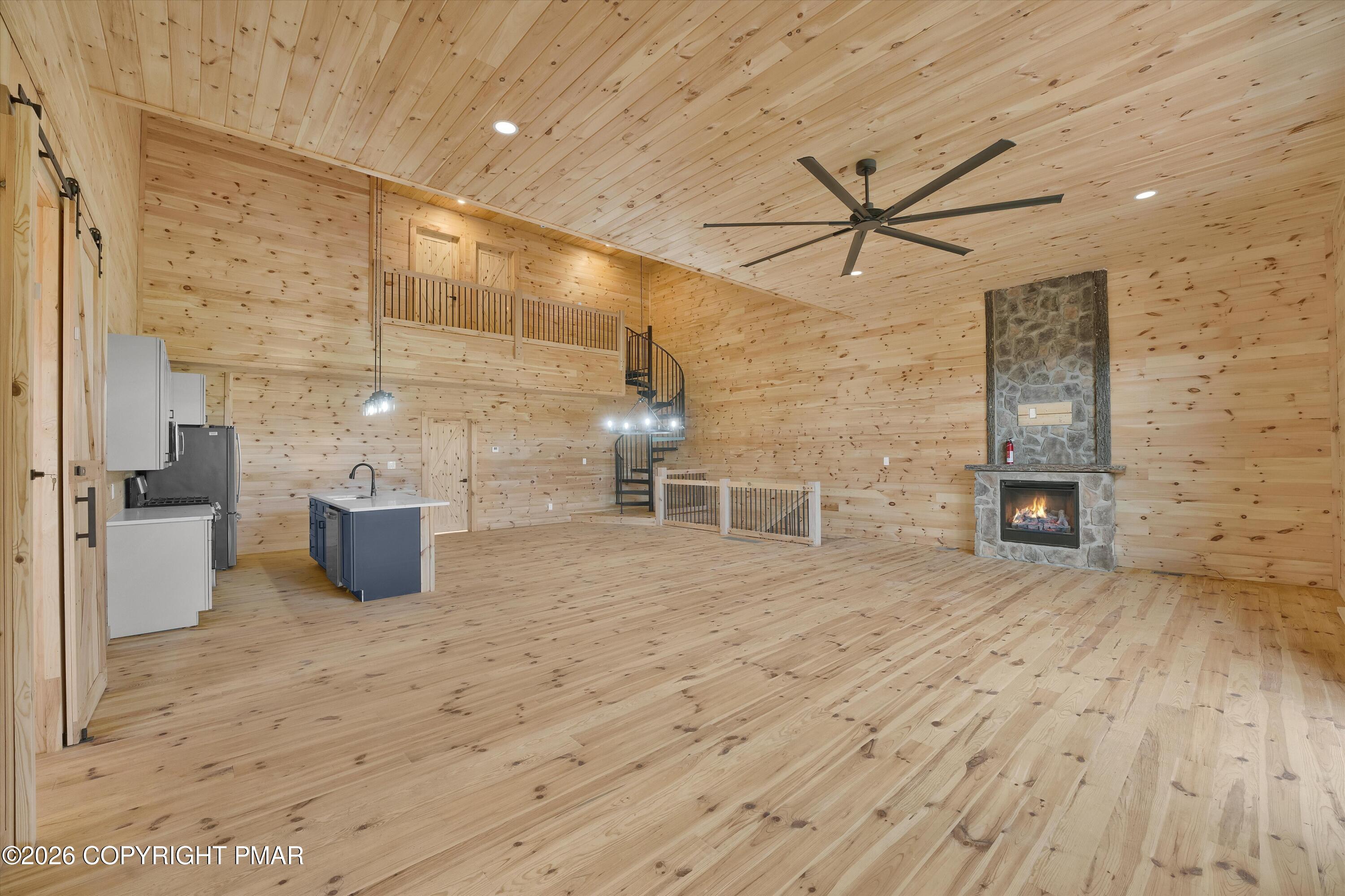 Wood-paneled open room with a loft staircase, kitchen island, and stone fireplace.