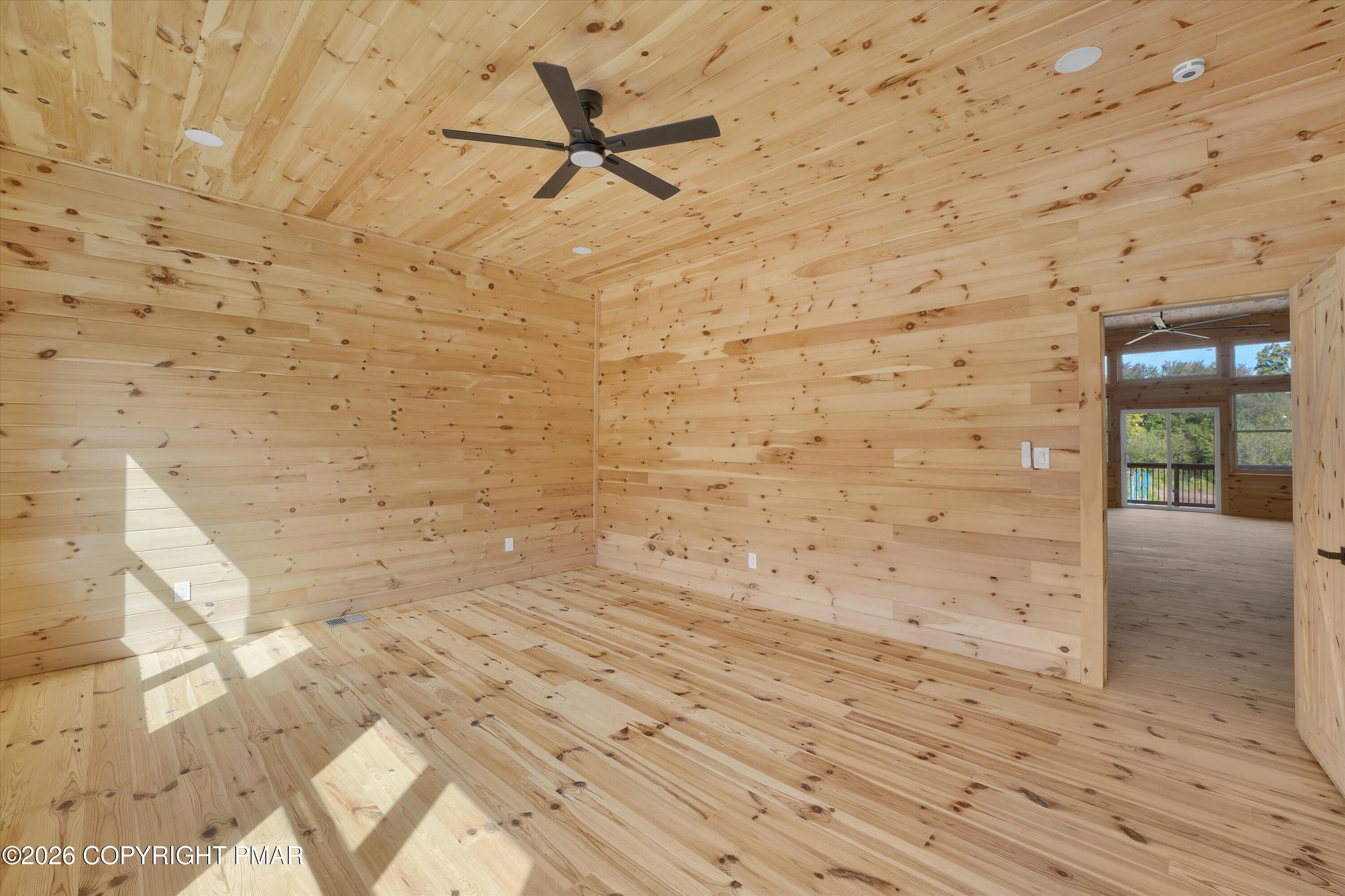 Empty knotty-pine room with wood-plank walls, ceiling, and floor, a ceiling fan, and doorway to a sunlit room.