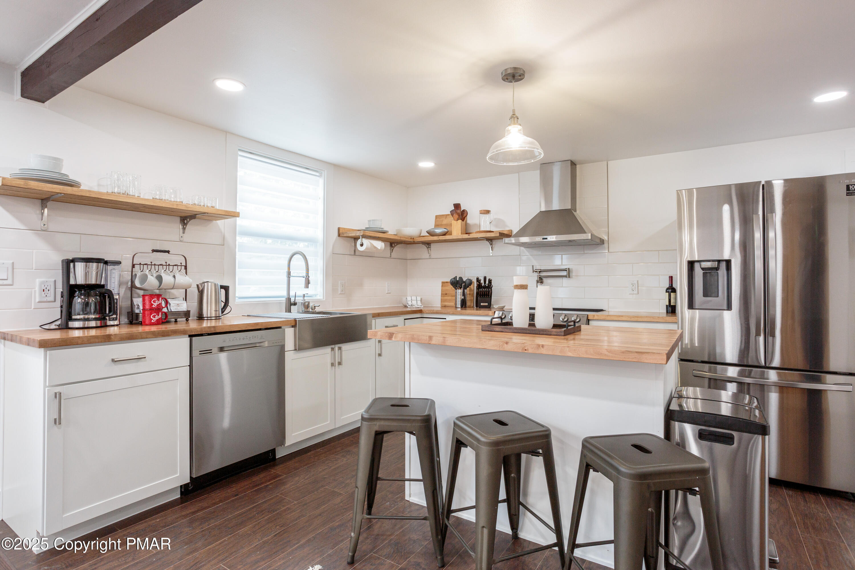 Modern kitchen with a wood-topped island, three metal stools, open shelving, and stainless steel appliances.
