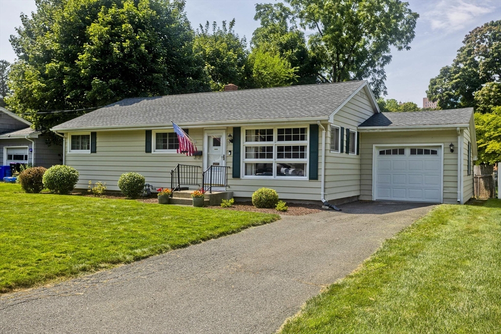 Single-story ranch home with green shutters, attached garage, American flag, and broad front lawn.