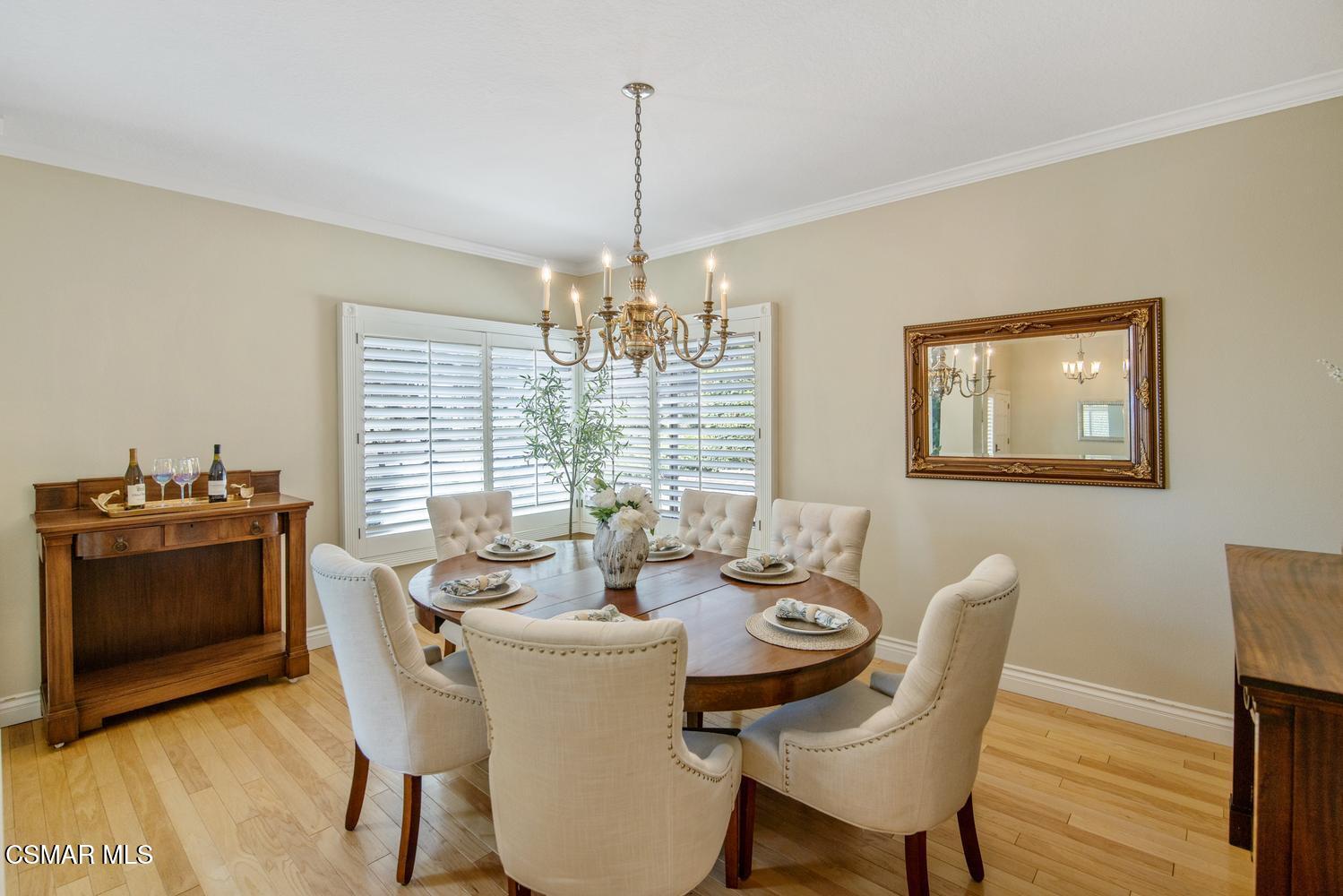 Dining room with a round wooden table, upholstered chairs, chandelier, and plantation shutters.