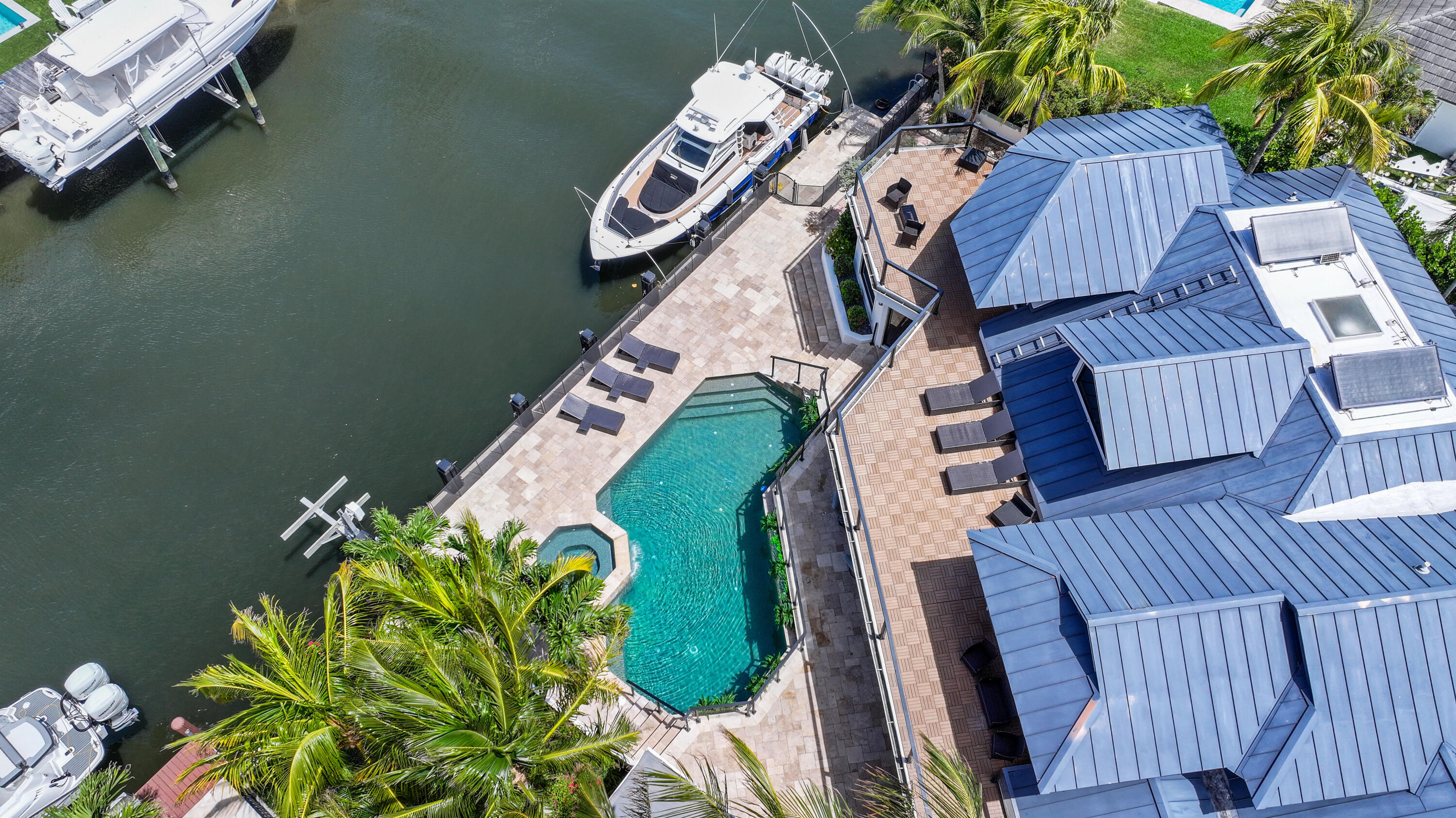 Waterfront home with a blue metal roof, curving pool, palm-lined patio, and boats on the canal.