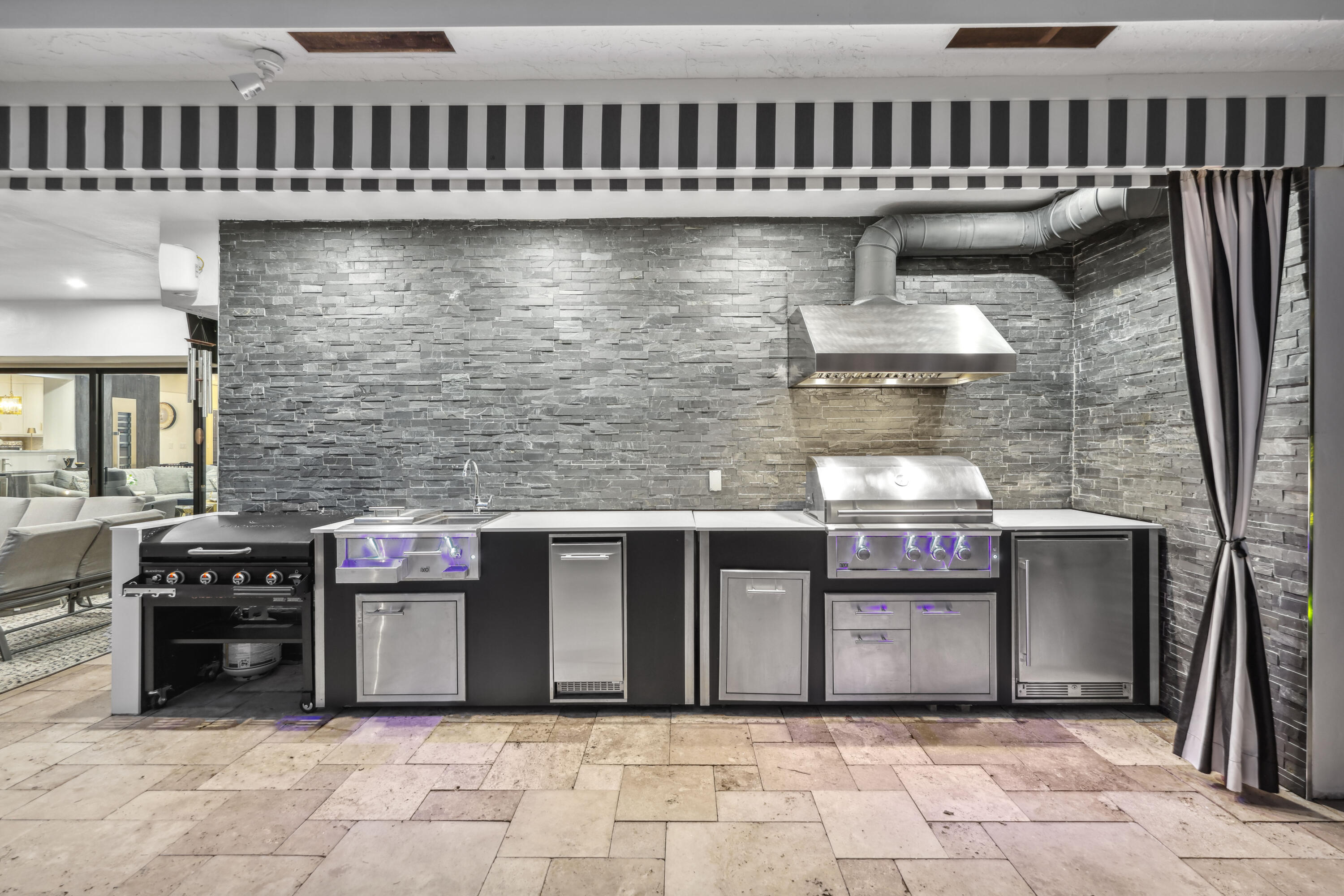 Outdoor kitchen with stainless steel grill and built-in appliances against a gray stone wall.