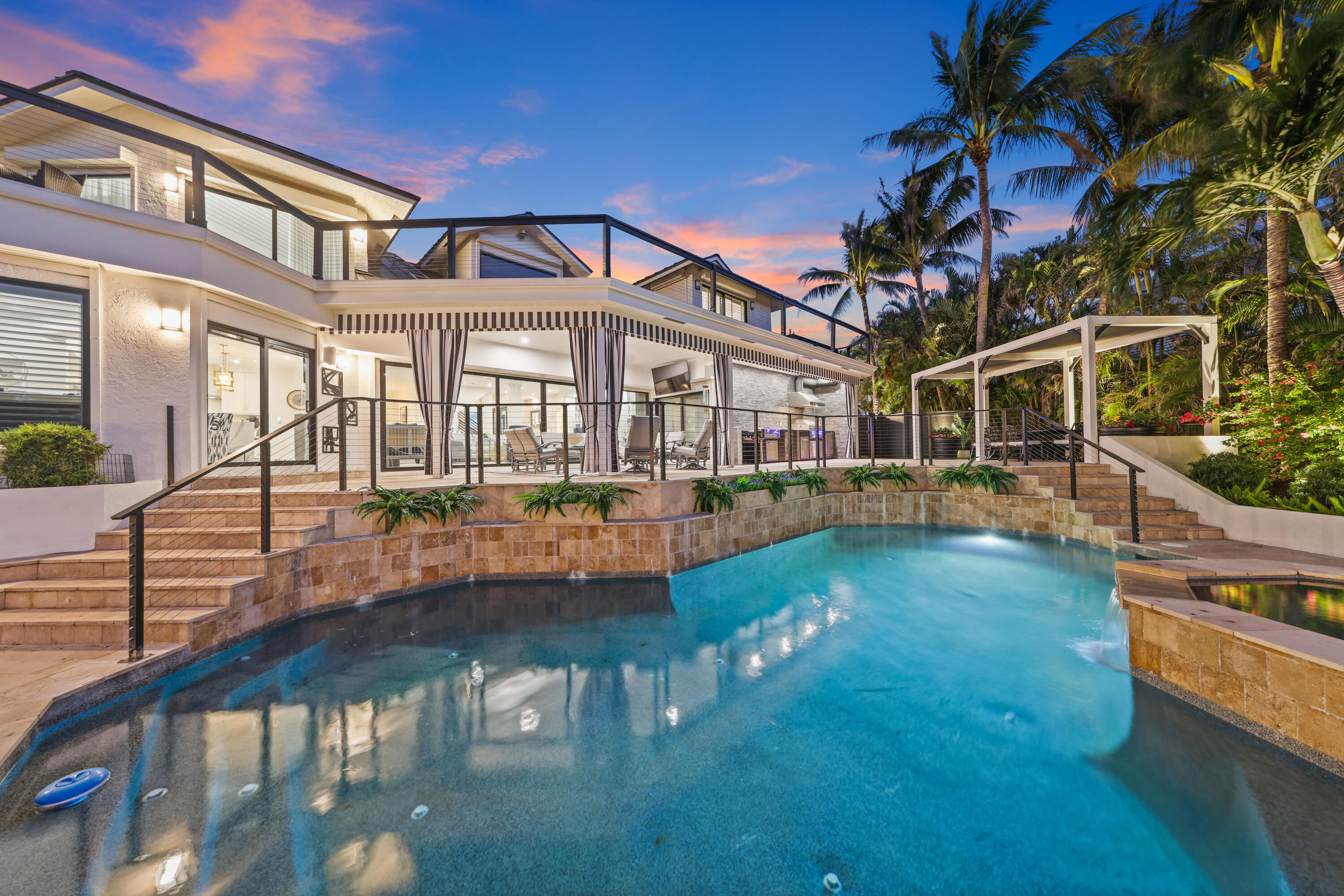 Modern home and illuminated pool at dusk, with a raised terrace and tropical palms.