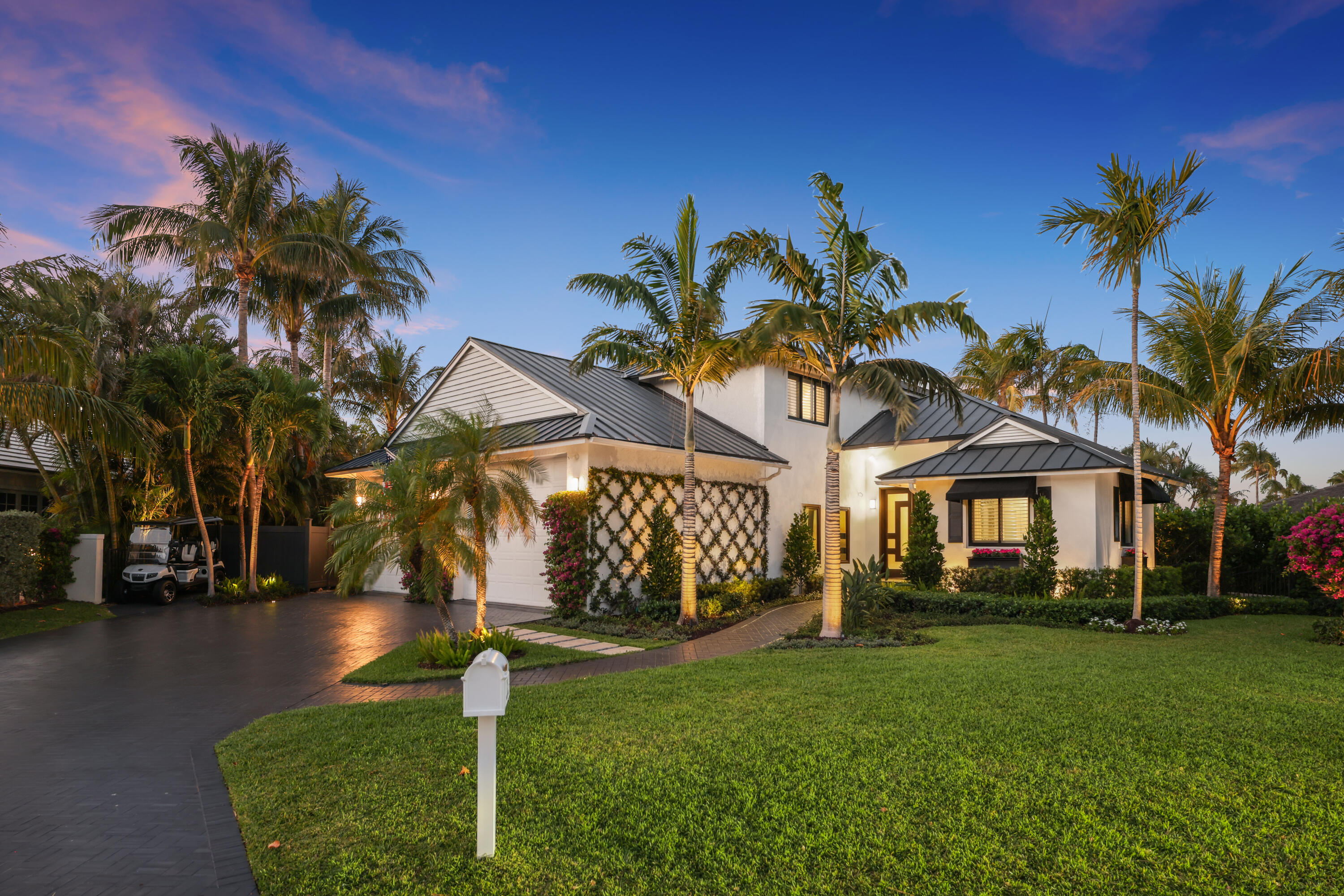 White tropical home with dark metal roofs, palm trees, and a broad lawn at dusk.
