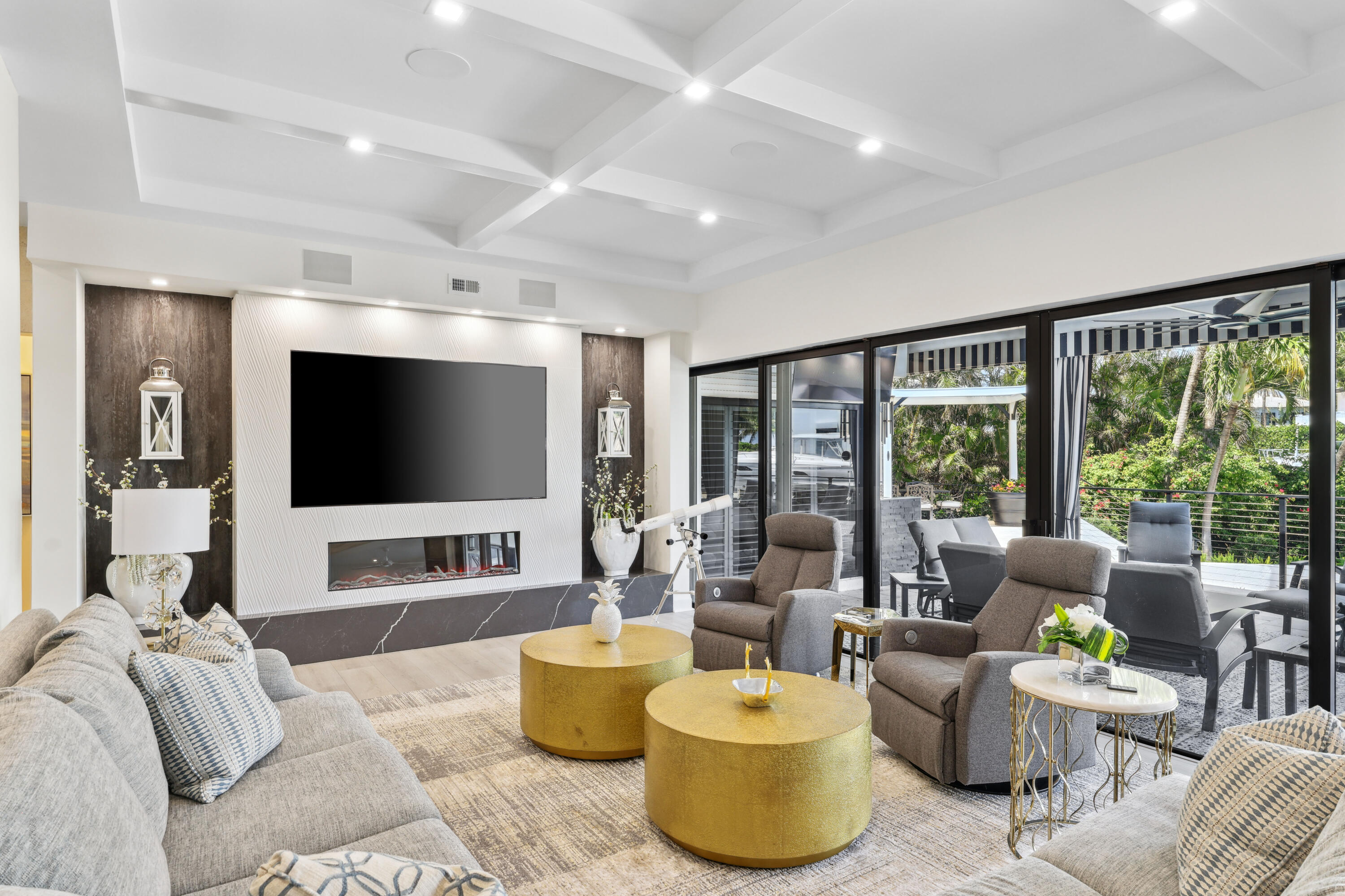 Living room with a coffered ceiling, linear fireplace, gold coffee tables, and glass doors to a tropical patio.