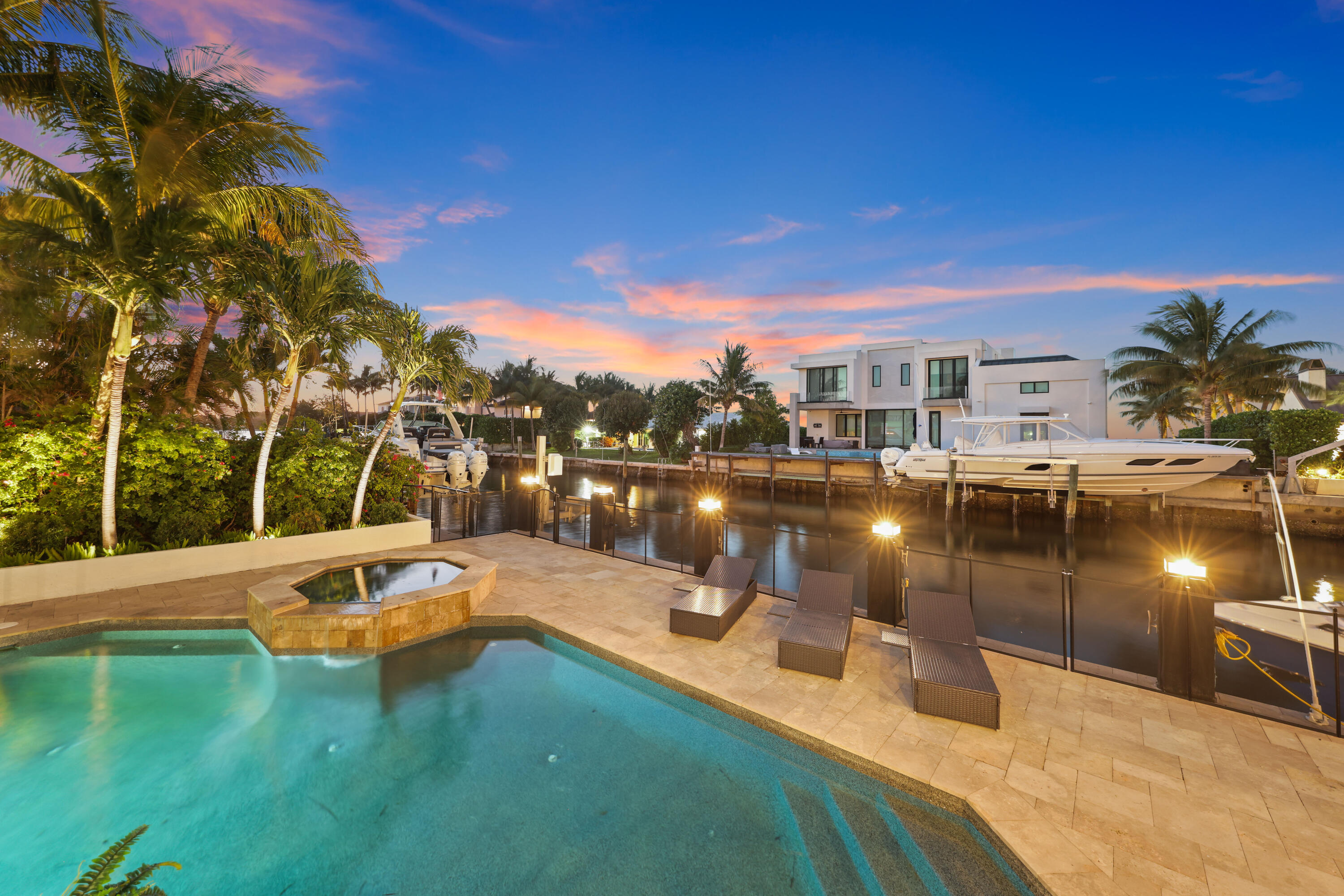 Modern waterfront home across a canal, with a pool, raised spa, docked boats, and palm-lined grounds at dusk.