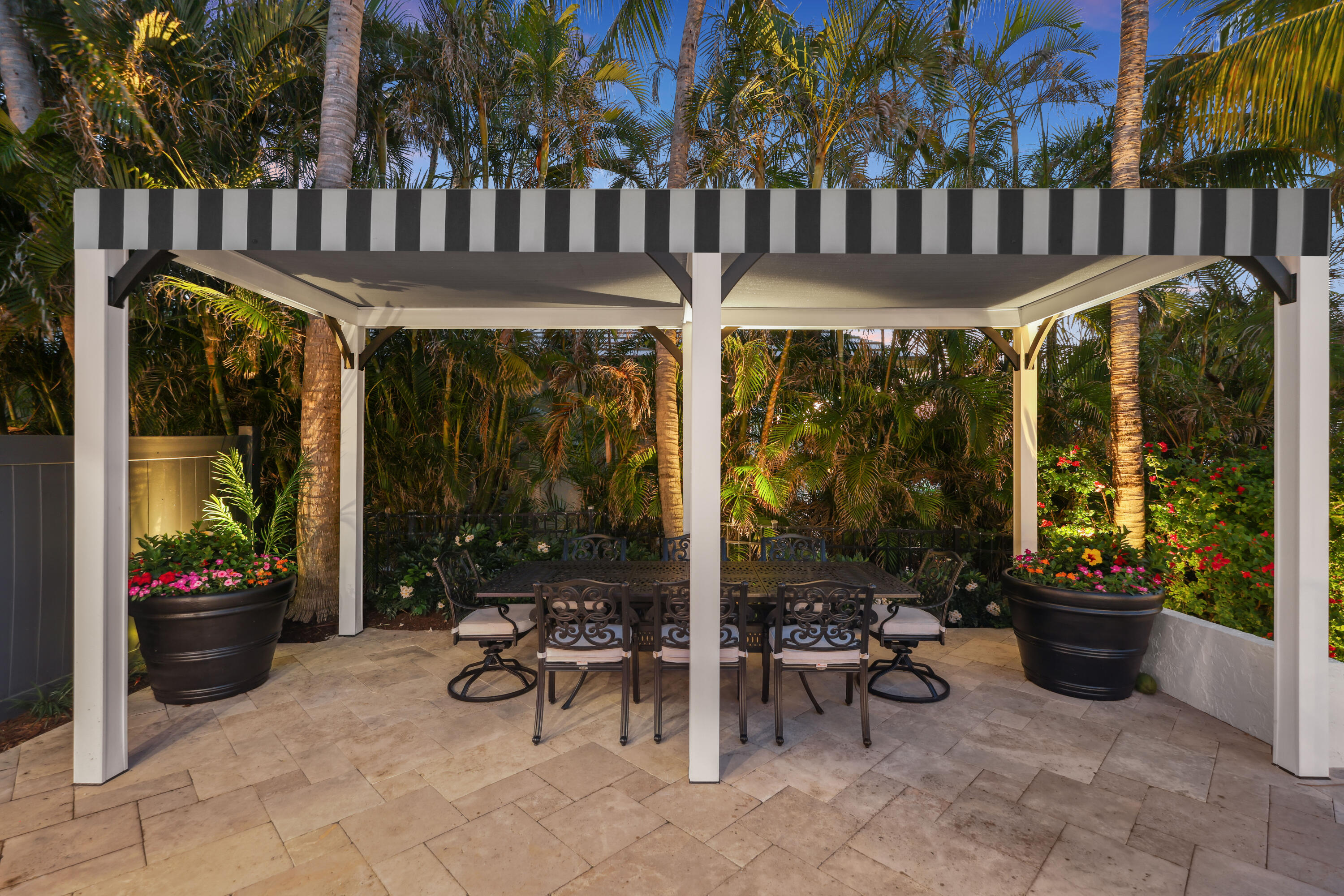 Covered patio dining area beneath a striped pergola, framed by tropical palms and flowering planters.