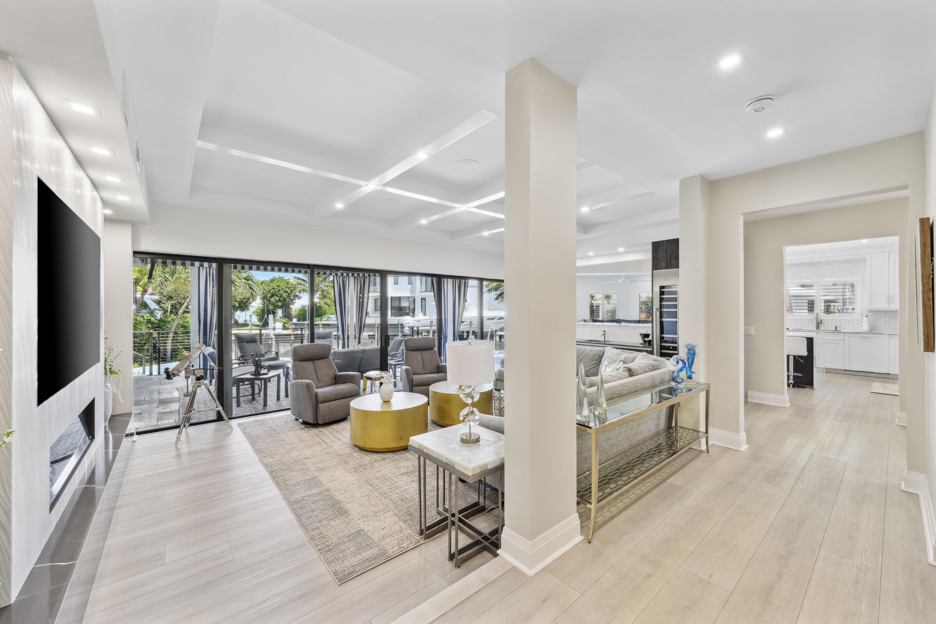 Open-plan living room with gray seating and gold tables, opening onto a patio and adjoining kitchen.