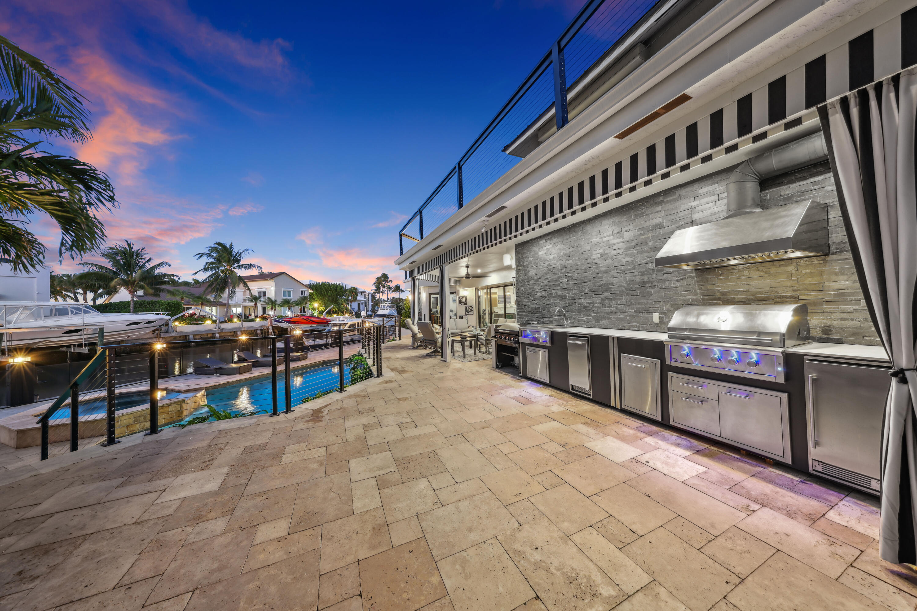 Waterfront patio with a built-in stainless steel outdoor kitchen, pool, and canal at dusk