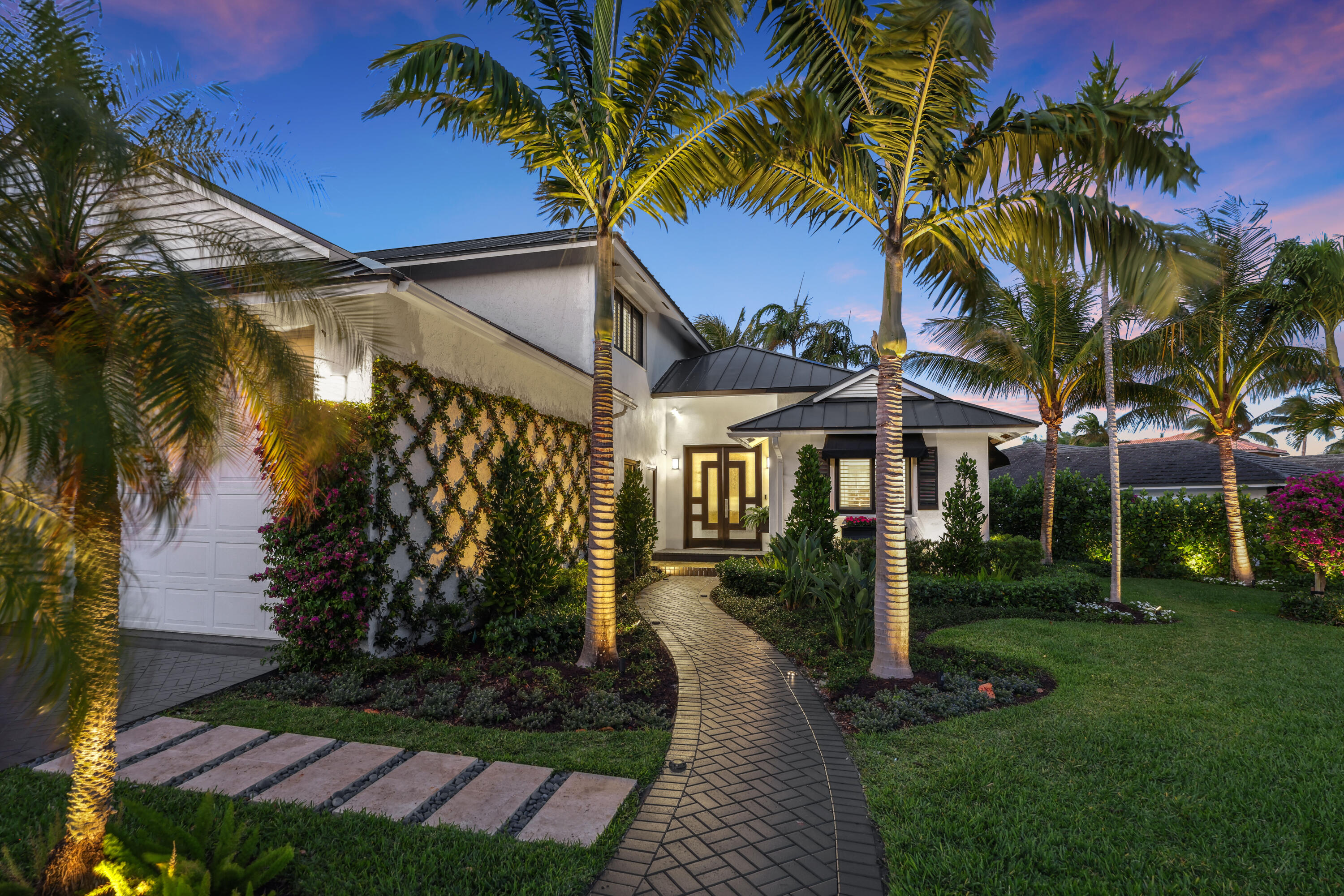 White two-story home with an illuminated entrance, curved walkway, and tall palm trees at dusk.