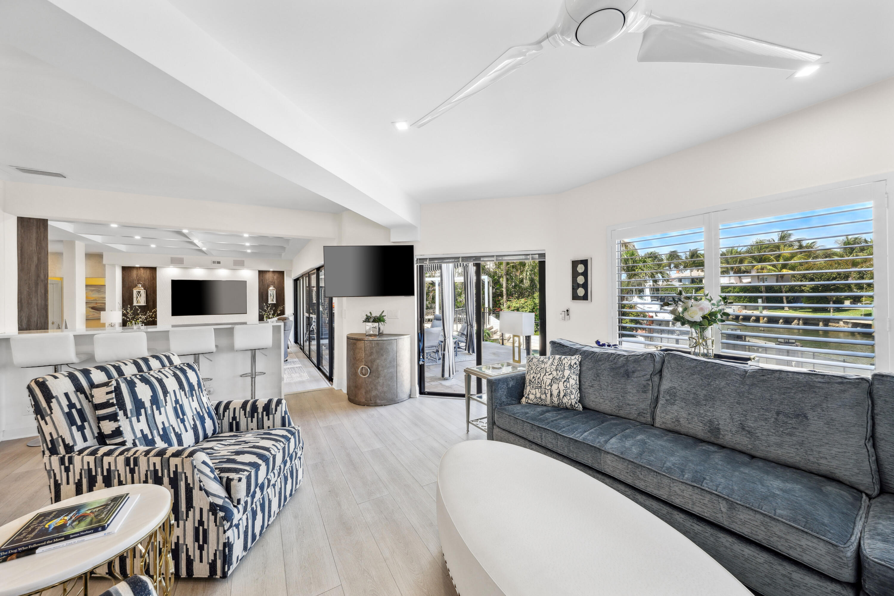 Open-plan living room with gray sofa, patterned armchair, and water views through wide louvered windows.