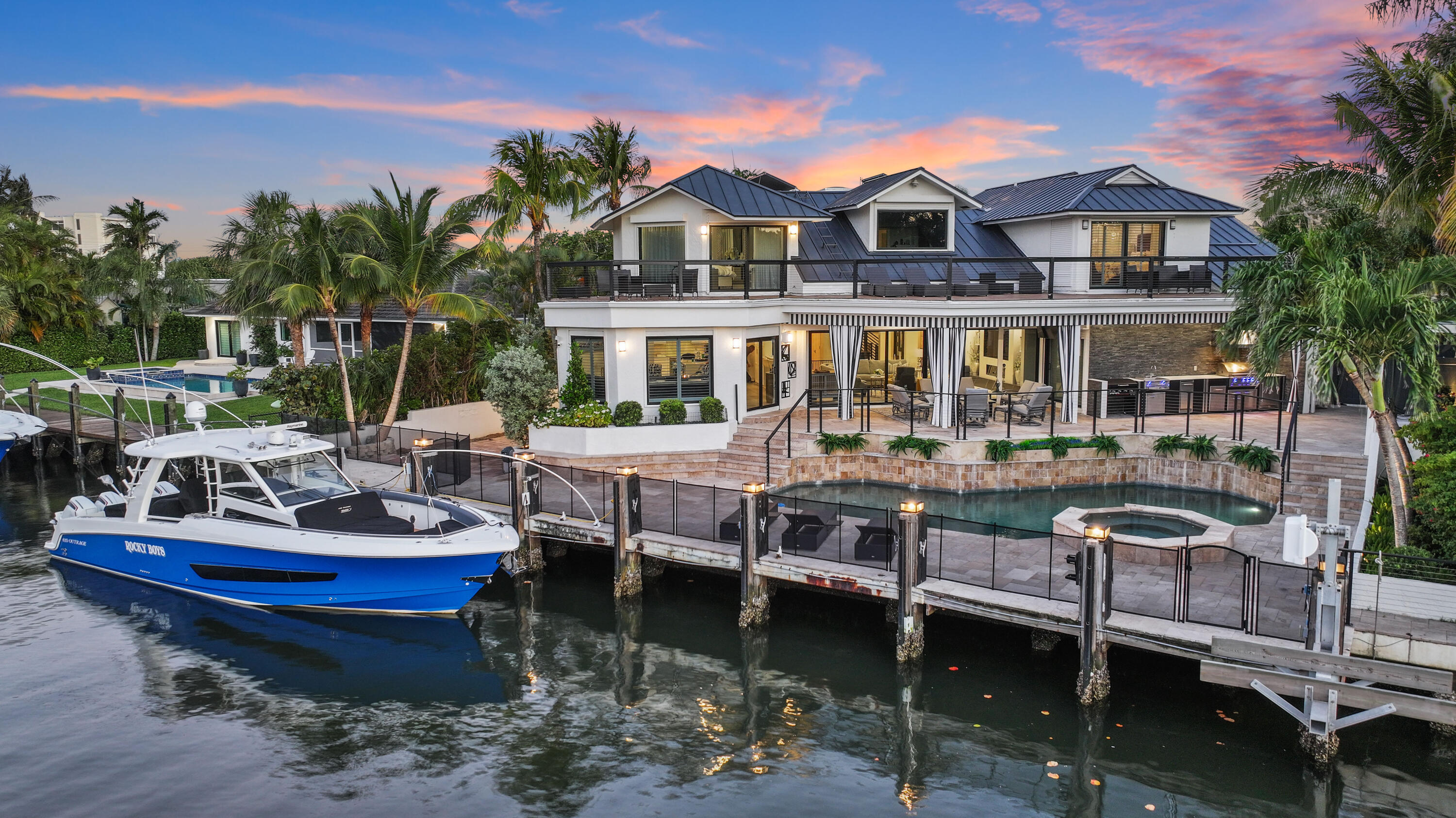 Waterfront home with a broad terrace, pool, spa, dock, and blue-and-white boat at dusk.