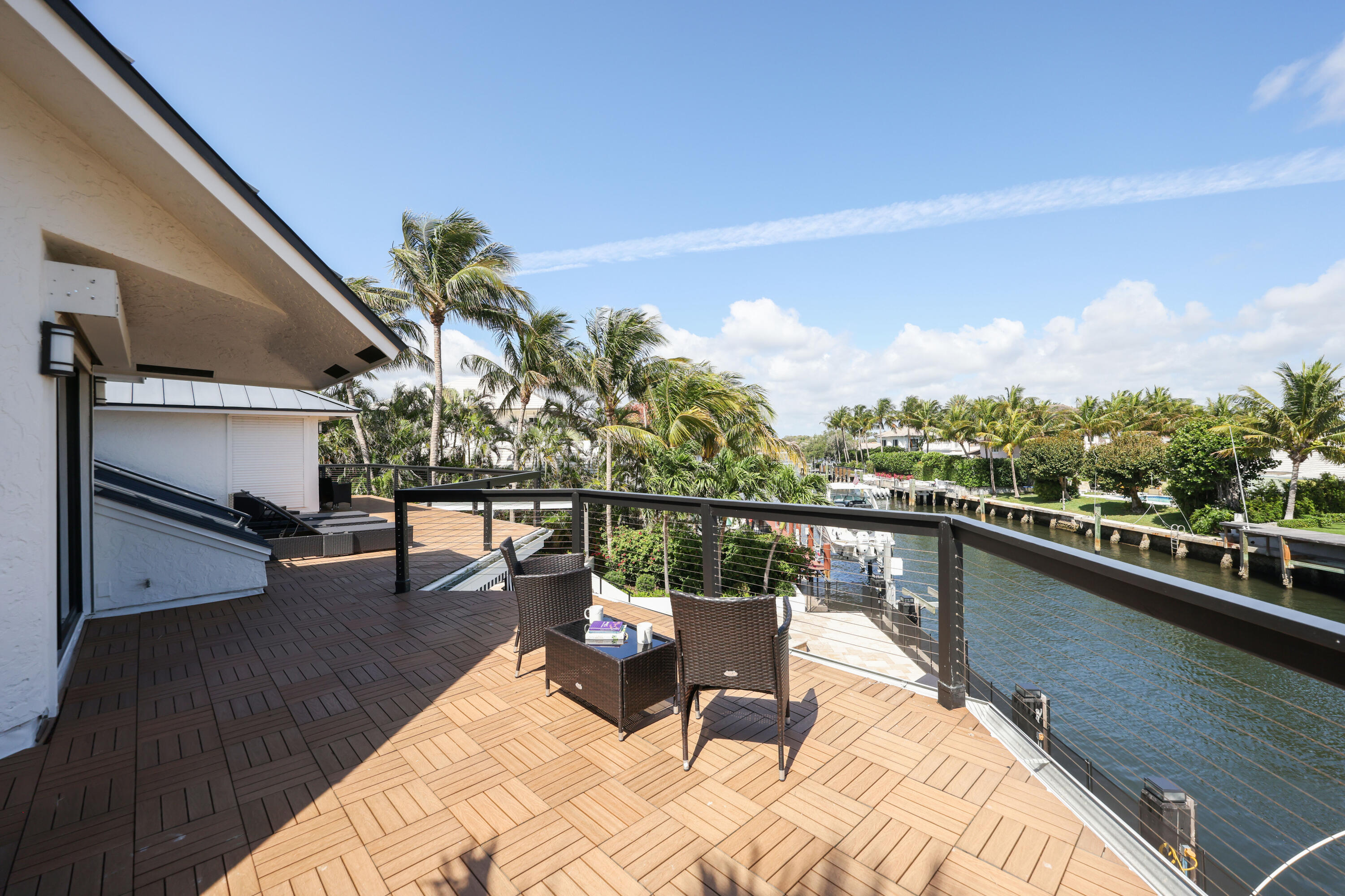 Furnished deck overlooking a palm-lined canal, with woven chairs and a cable railing.