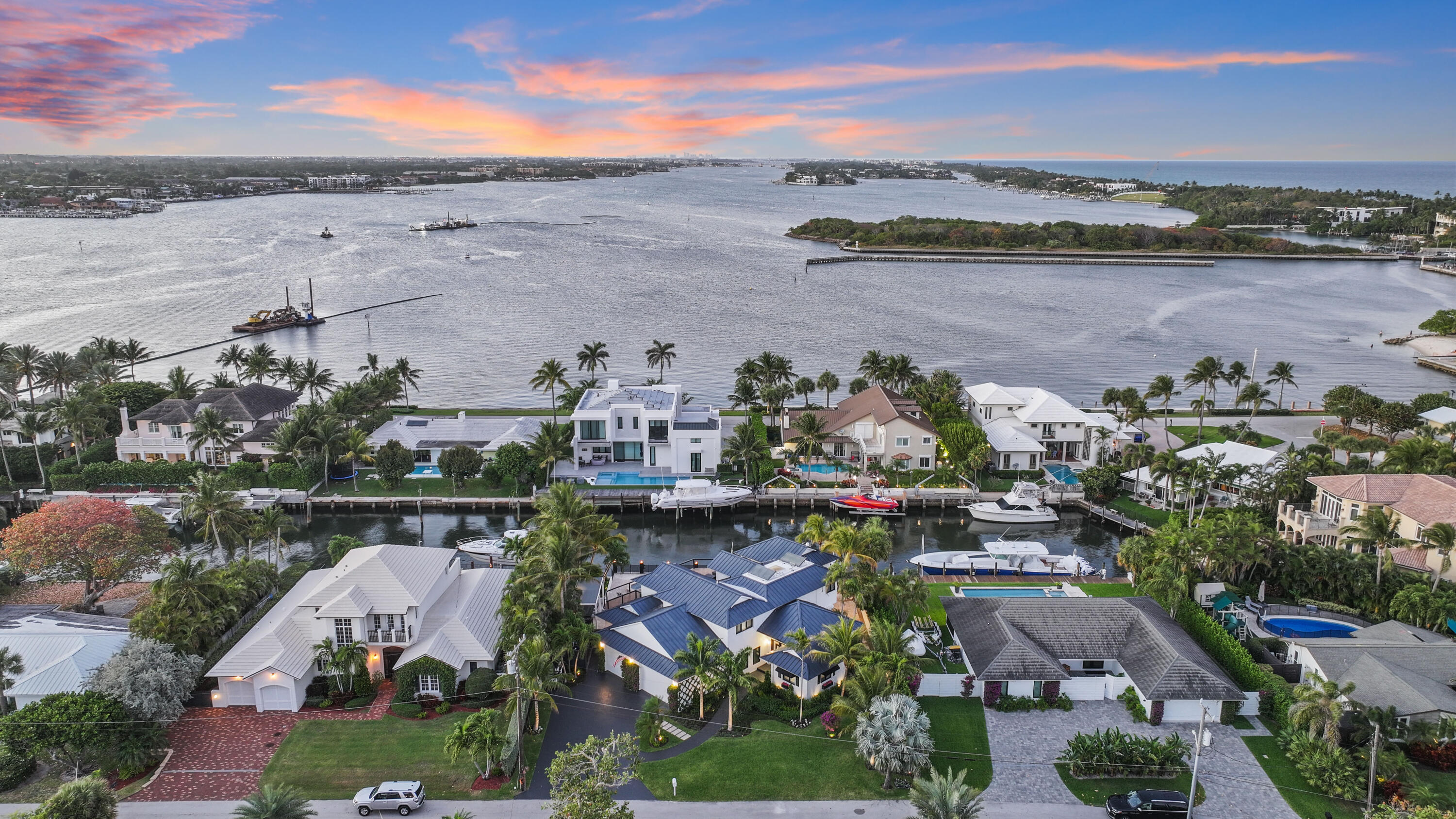 Aerial view of a modern waterfront home, neighboring docks and boats, and a wide waterway at dusk.