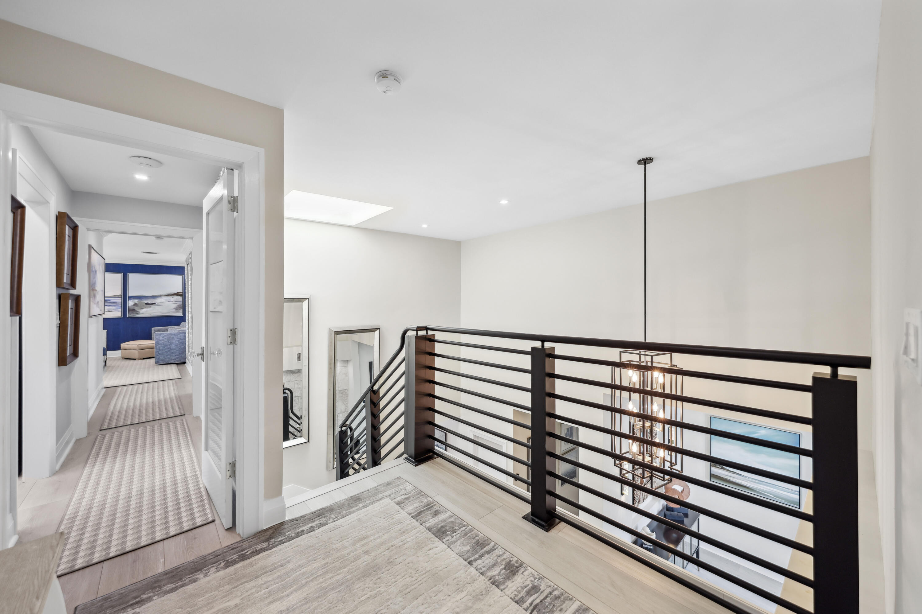 Upstairs landing with black railing overlooking a chandelier and a hallway leading to a blue-accented sitting room.