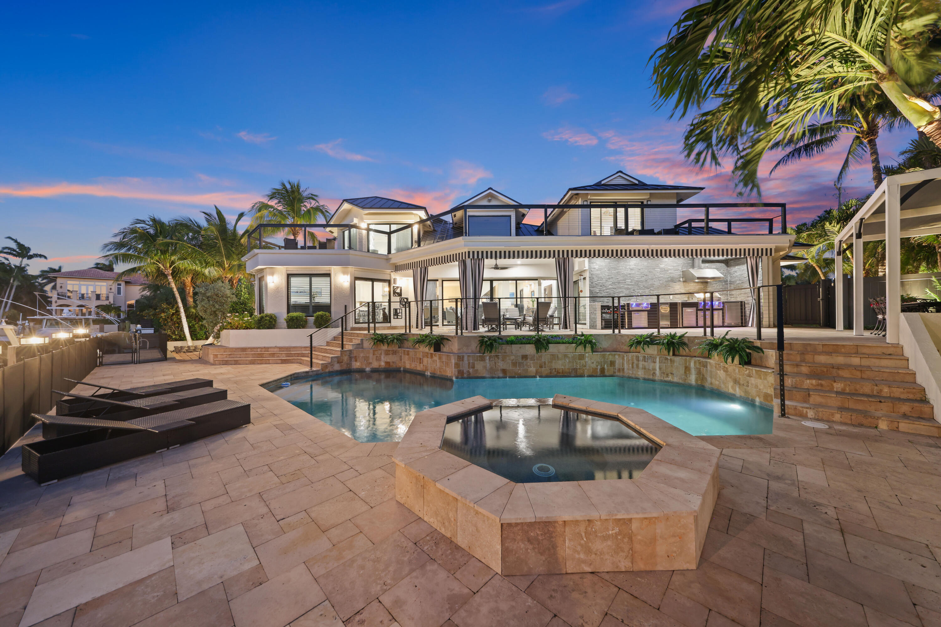 Luxury home’s illuminated rear exterior with a swimming pool, raised spa, and palm-lined patio at dusk.