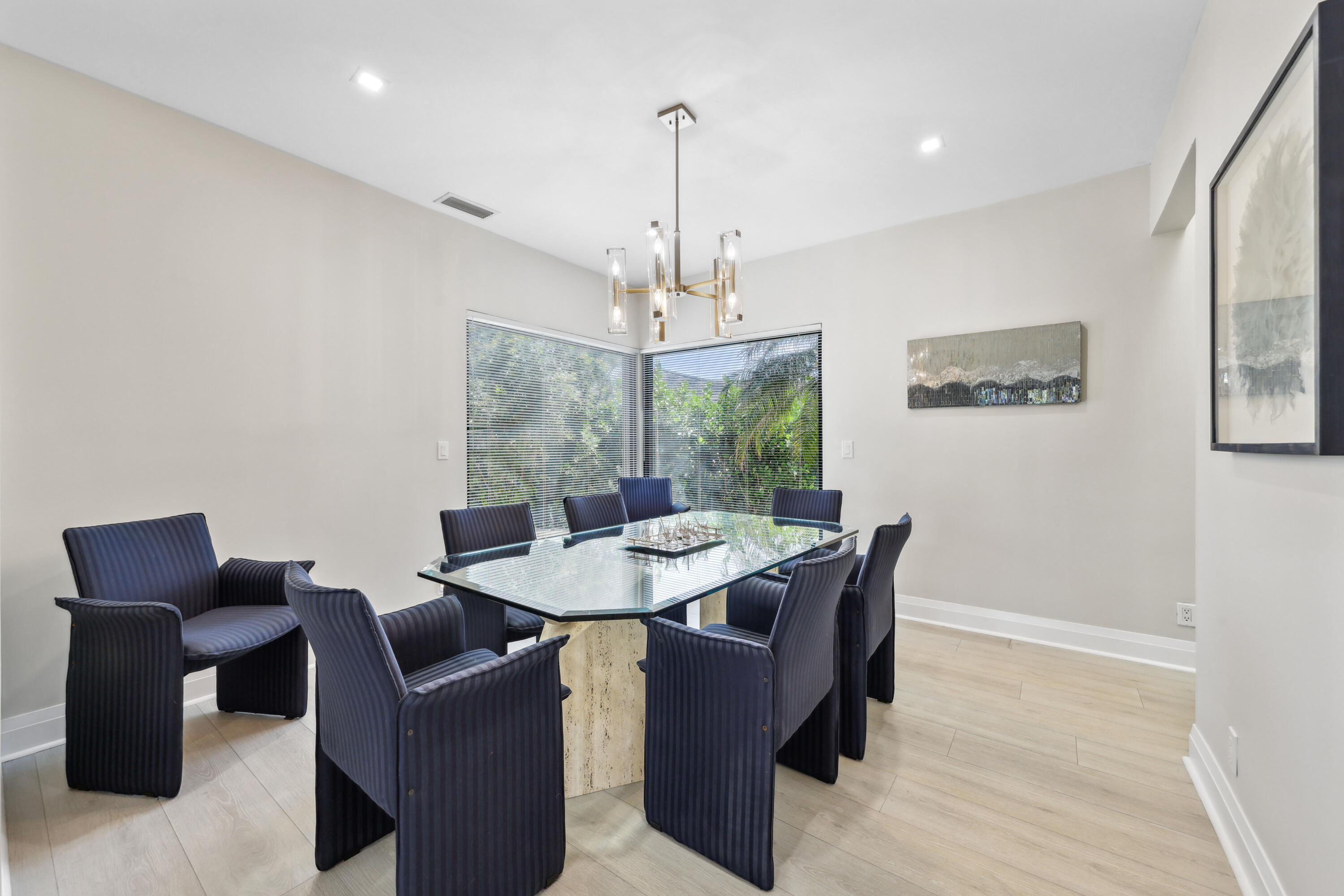 Dining room with a glass-top table, dark chairs, and corner windows overlooking greenery.