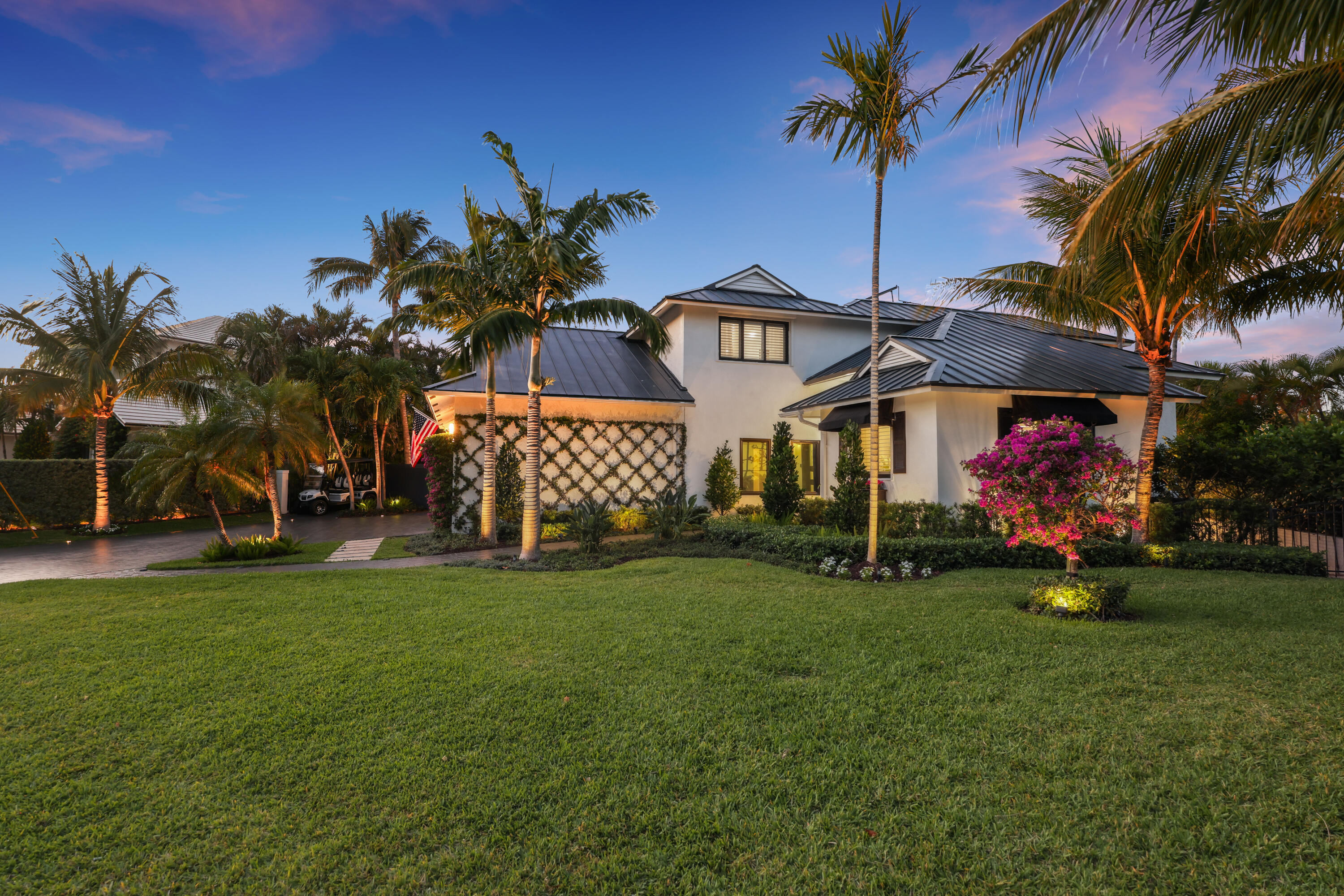 White two-story home with a dark metal roof, palm trees, and tropical landscaping.