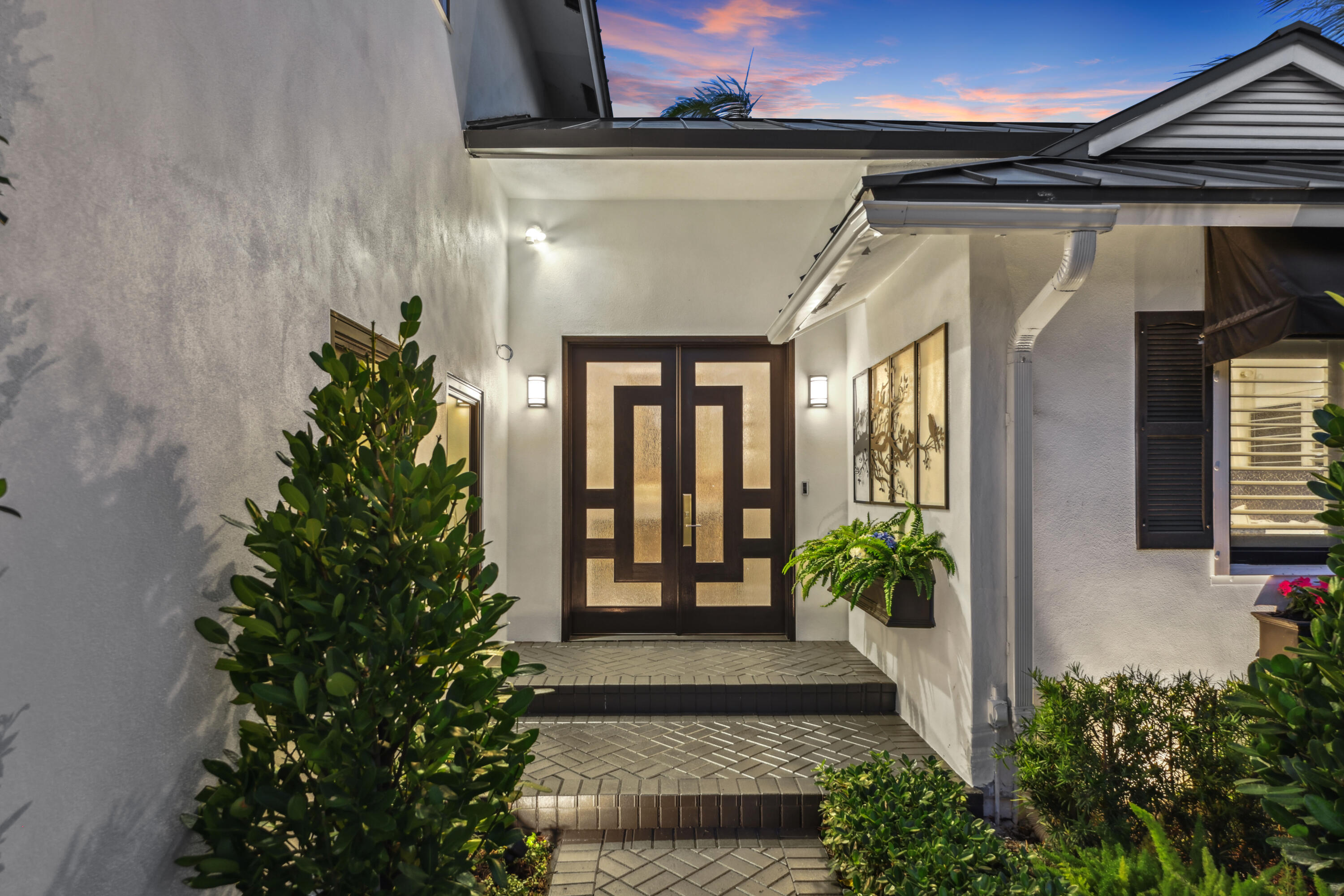 Home entrance with patterned double doors, tiled steps, lush plants, and a pink dusk sky.
