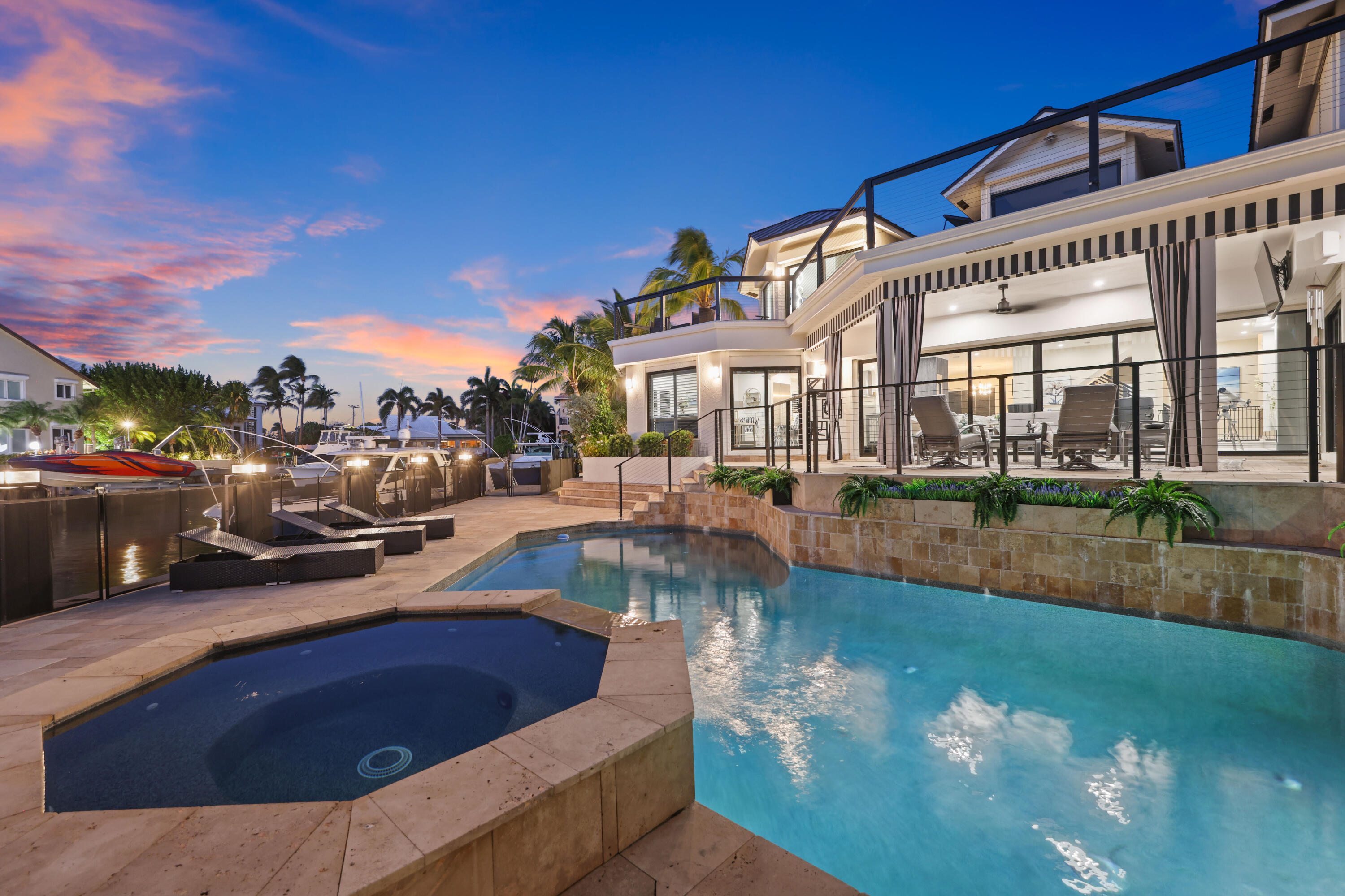 Waterfront home with illuminated pool and spa, patio, and boats along the canal at dusk.