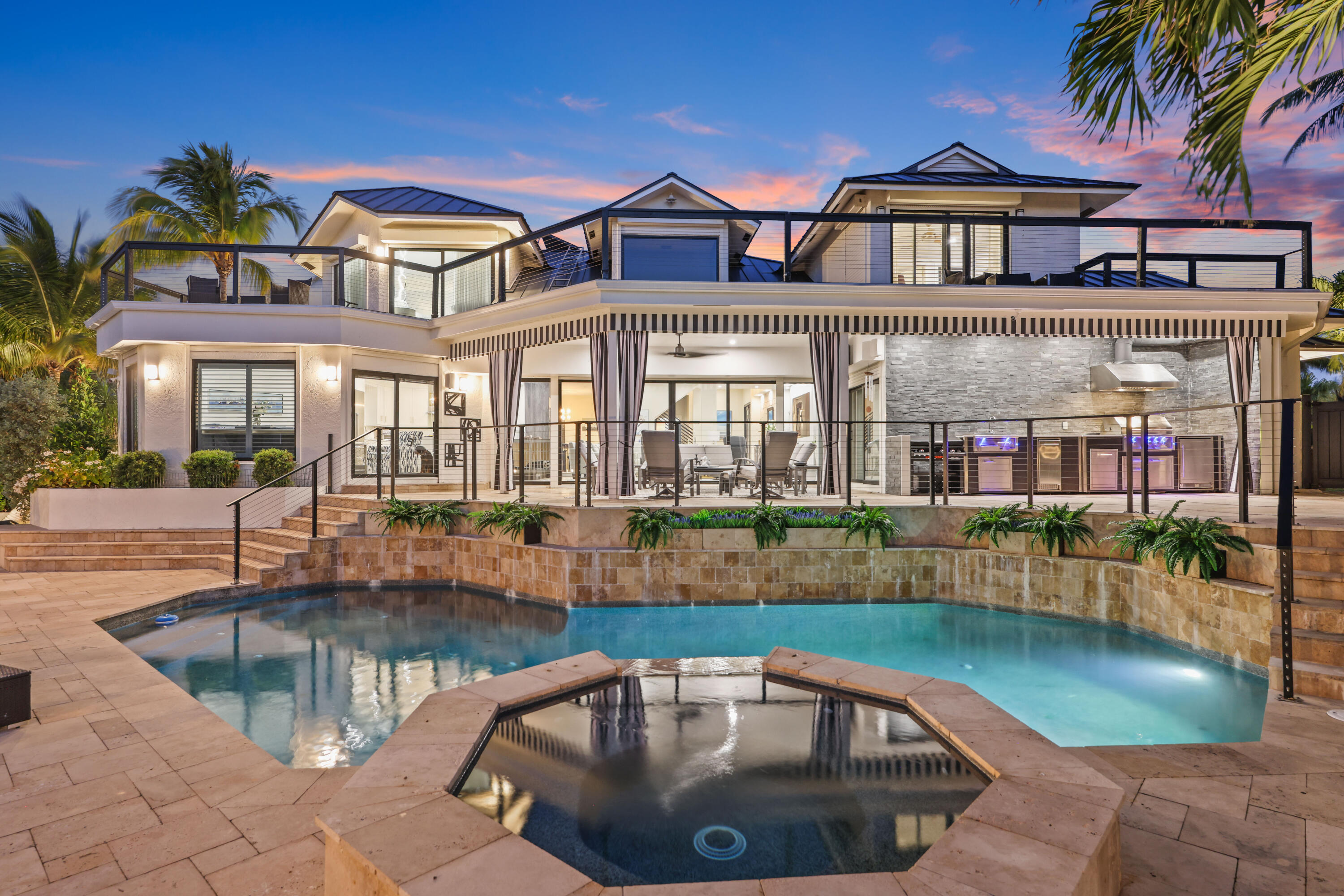 Modern tropical home with a glass-railed terrace, outdoor kitchen, pool, and spa at dusk