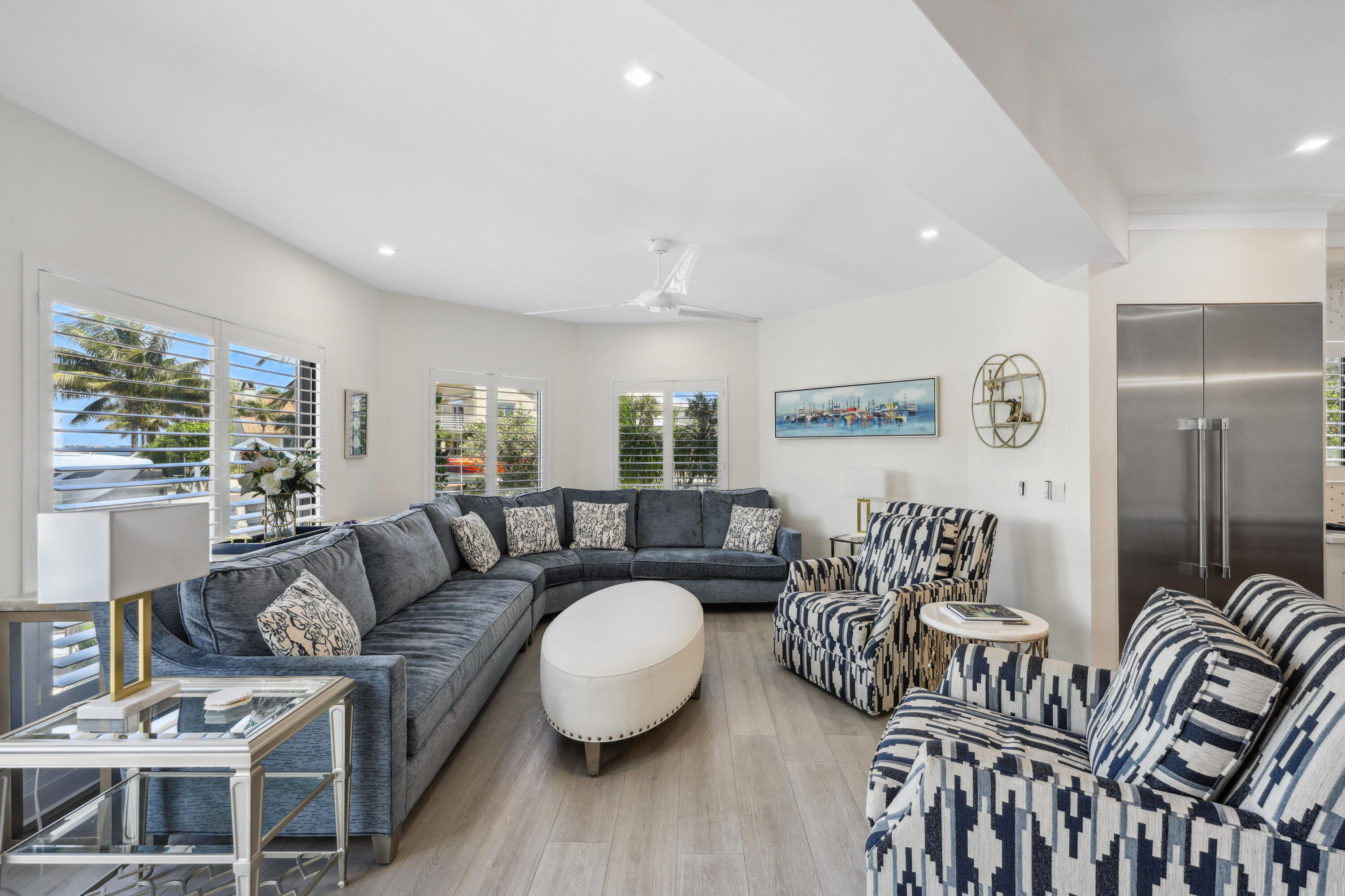 Furnished living room with a gray sectional, patterned armchairs, and plantation shutters overlooking palm trees.