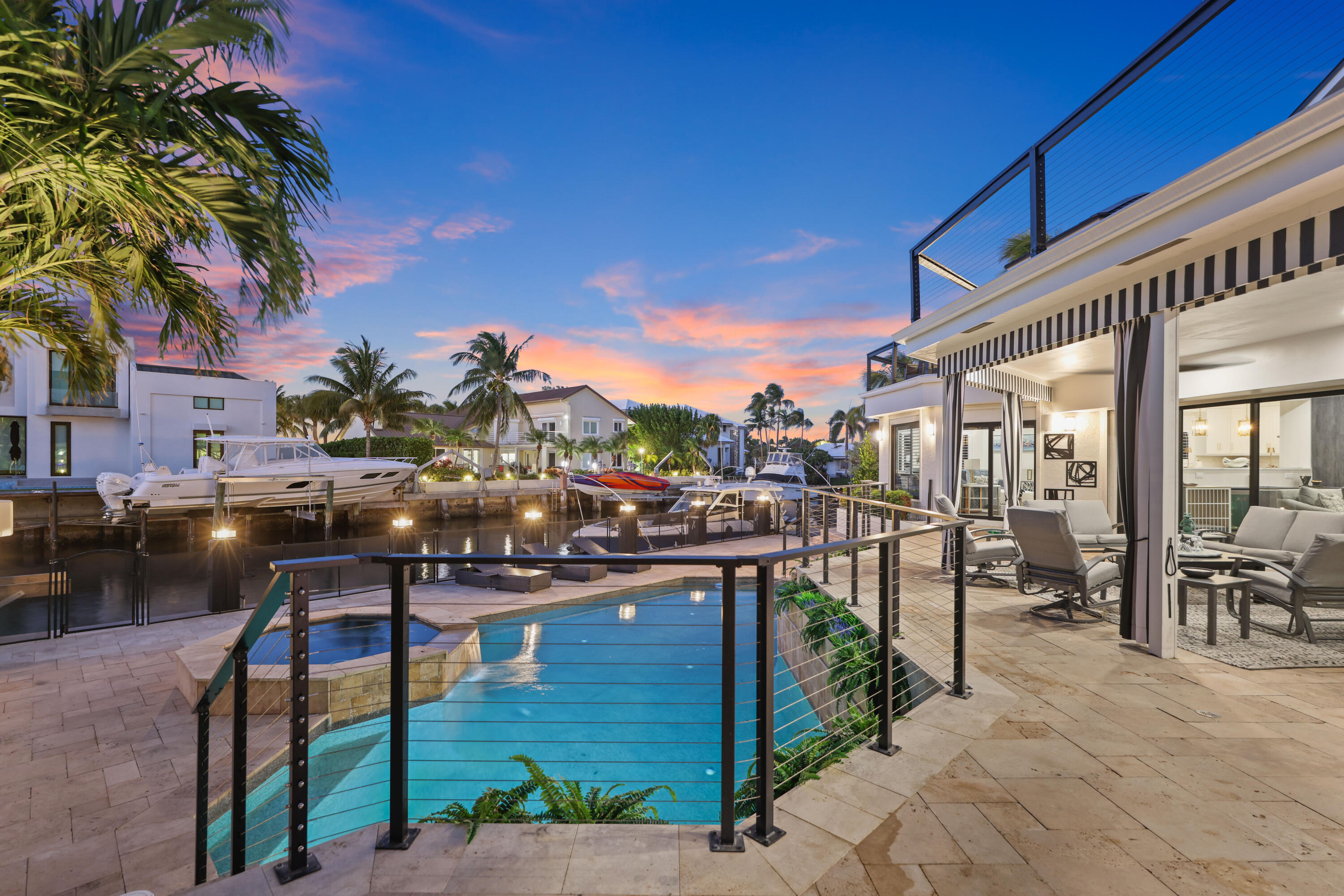 Waterfront home with a pool and furnished patio overlooking a boat-lined canal at sunset.