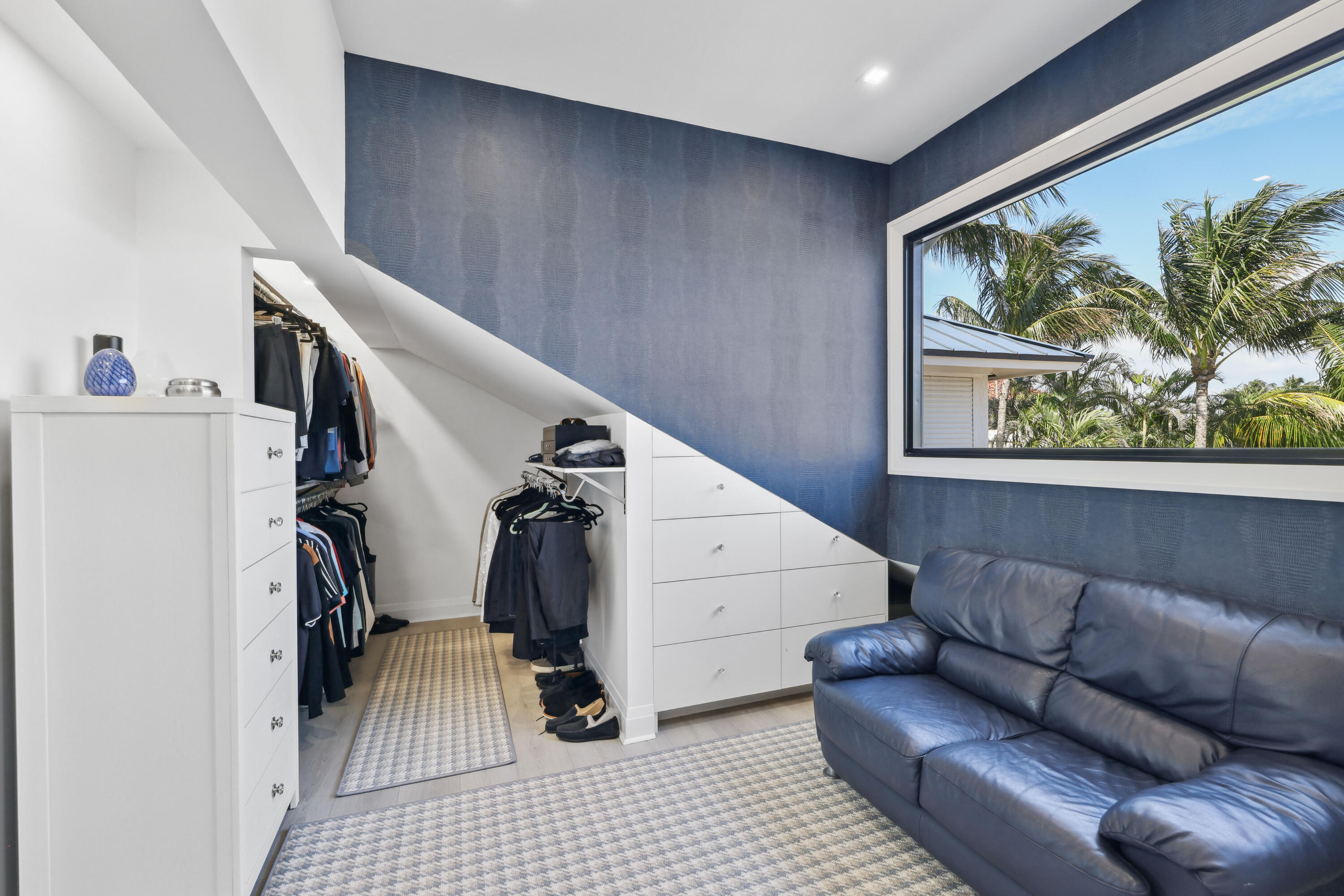 Walk-in dressing room with sloped built-in drawers, clothing rails, and a blue leather sofa by a palm-framed window.