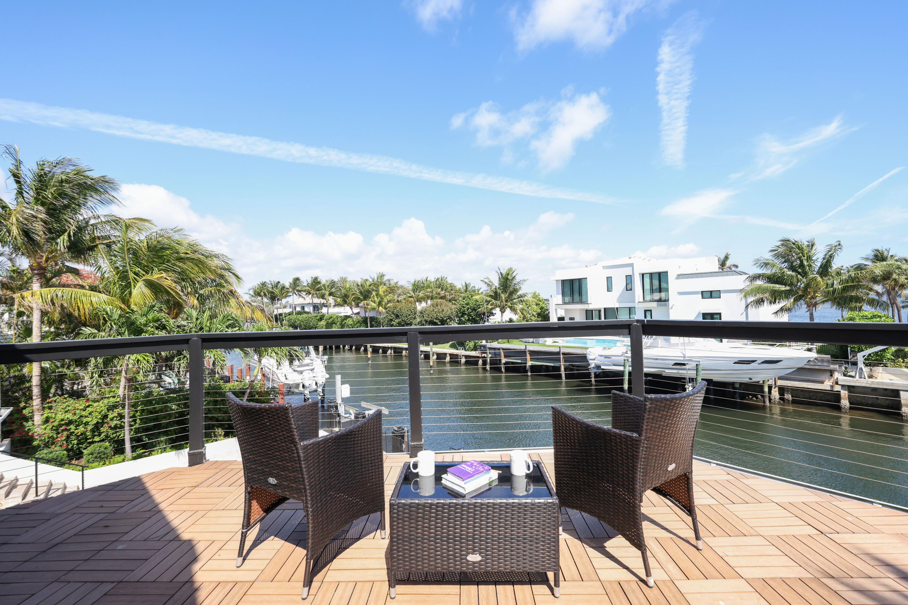 Waterfront deck with wicker seating overlooking a palm-lined canal and docked boats.