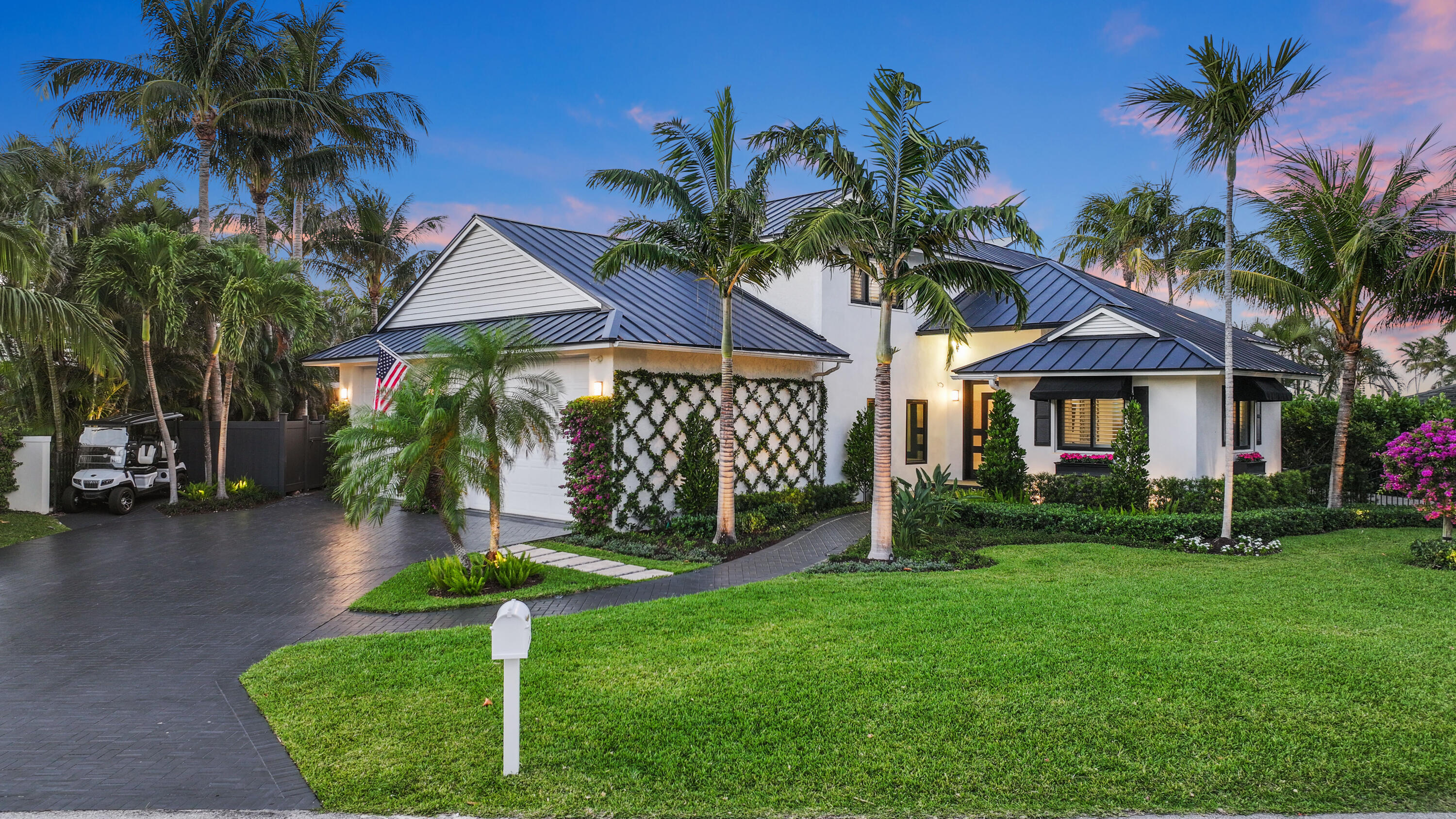 White tropical home with dark metal roofs, palm trees, and a curved paved driveway at dusk.