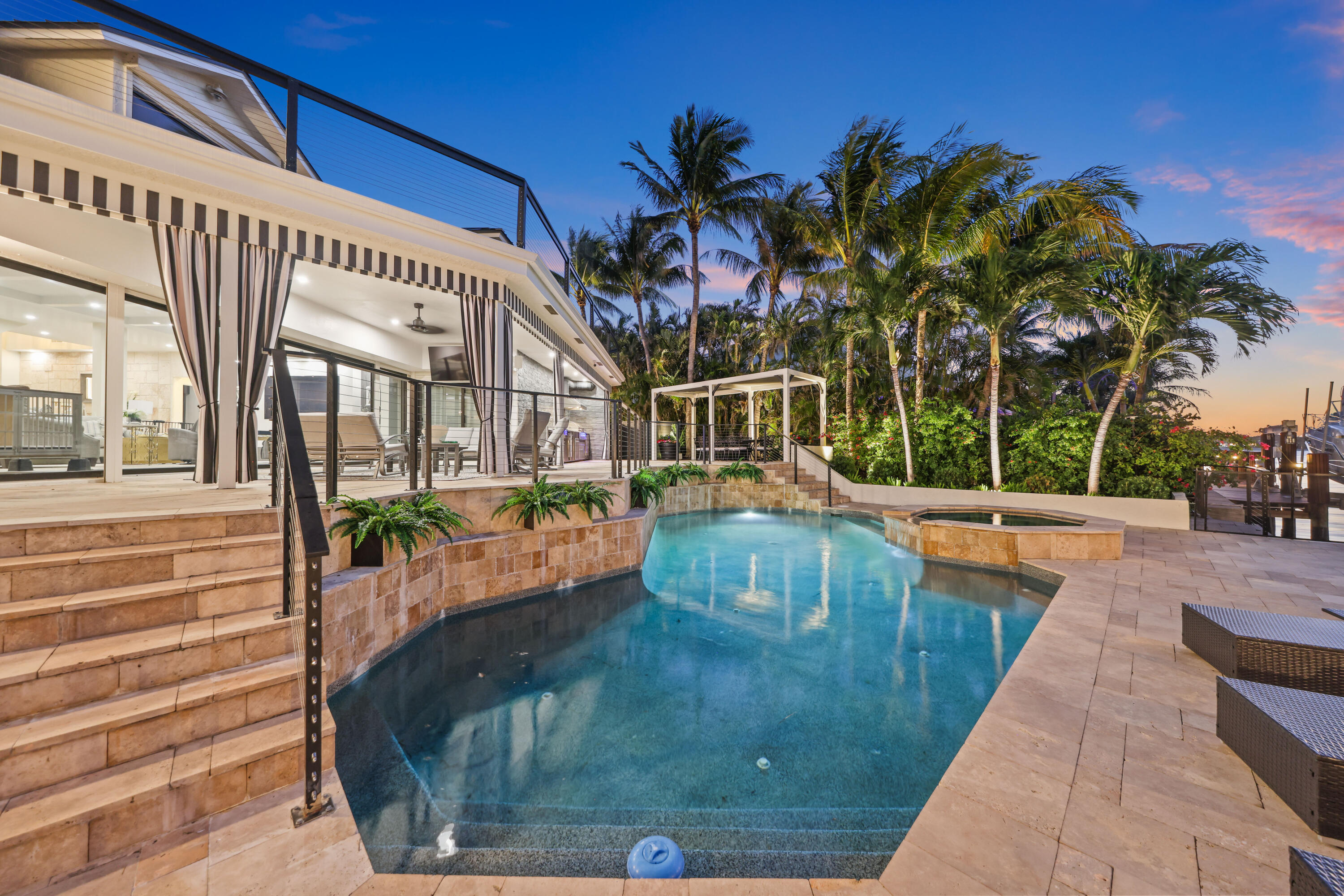 Backyard swimming pool and raised spa beside a stone patio and pergola at dusk.