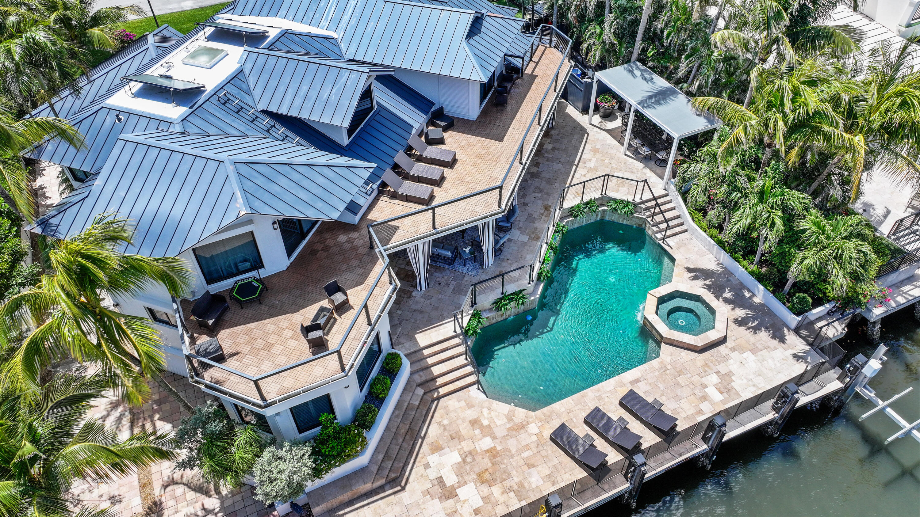 Aerial view of a waterfront home with a turquoise pool, spa, and furnished terraces.