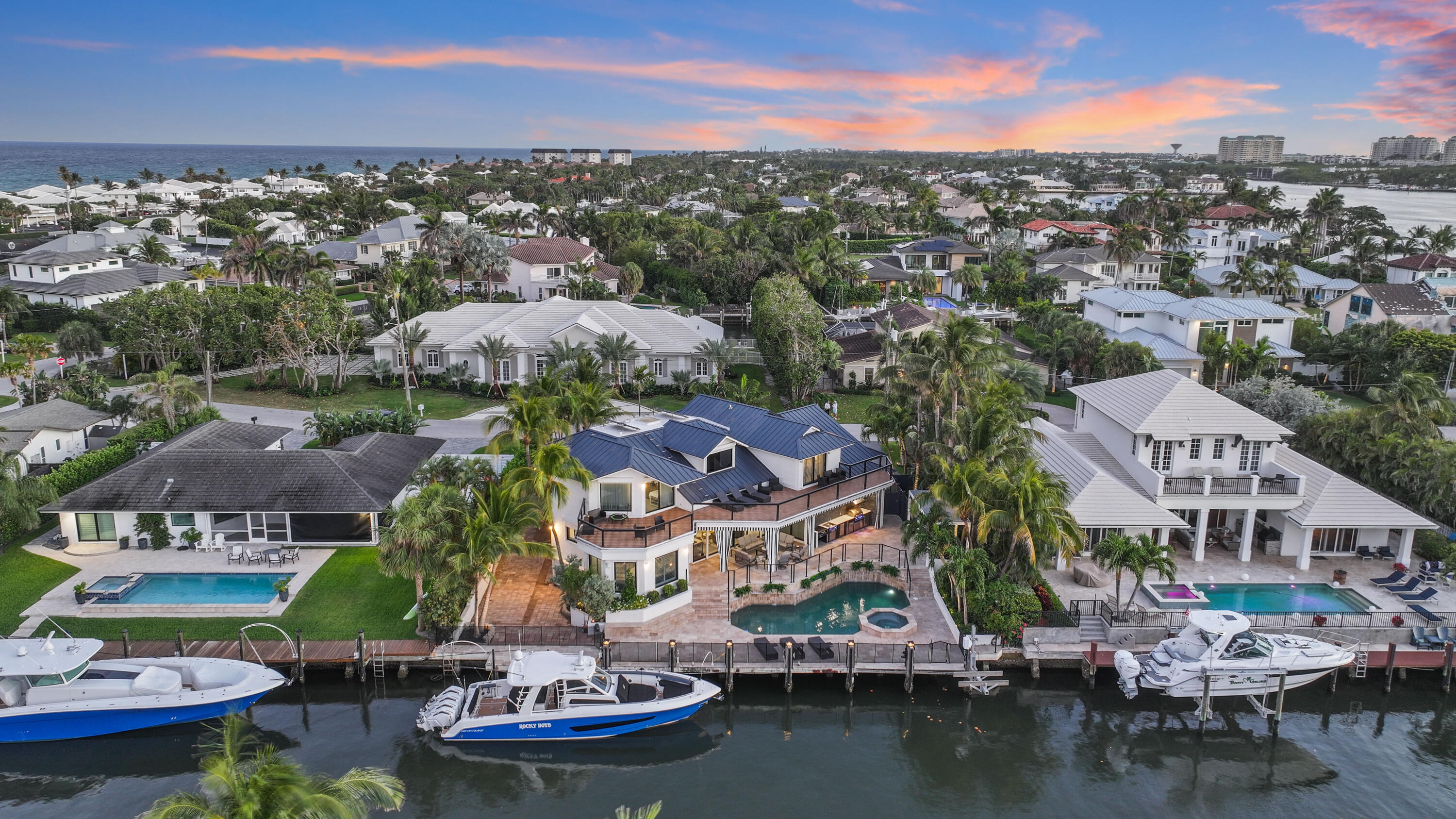 Waterfront home with a blue roof, pool, and docked boats, with the ocean beyond.
