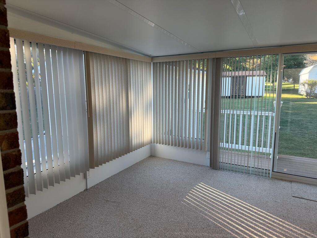 Empty carpeted sunroom with vertical blinds and sliding glass doors opening toward a deck and lawn.