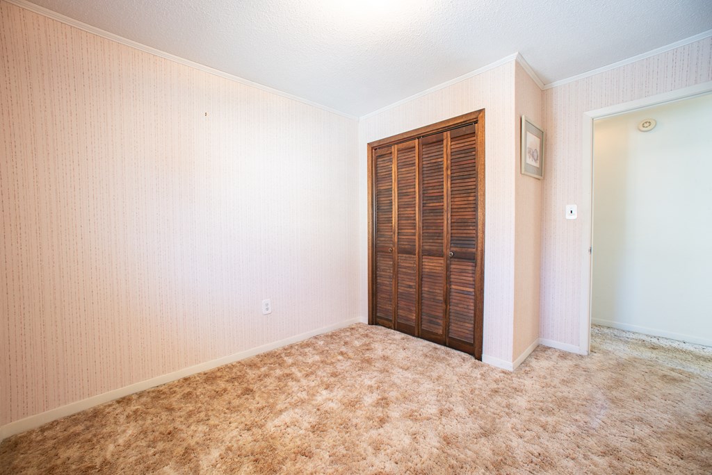 Empty bedroom with tan carpet, pink patterned wallpaper, and dark wood louvered closet doors.