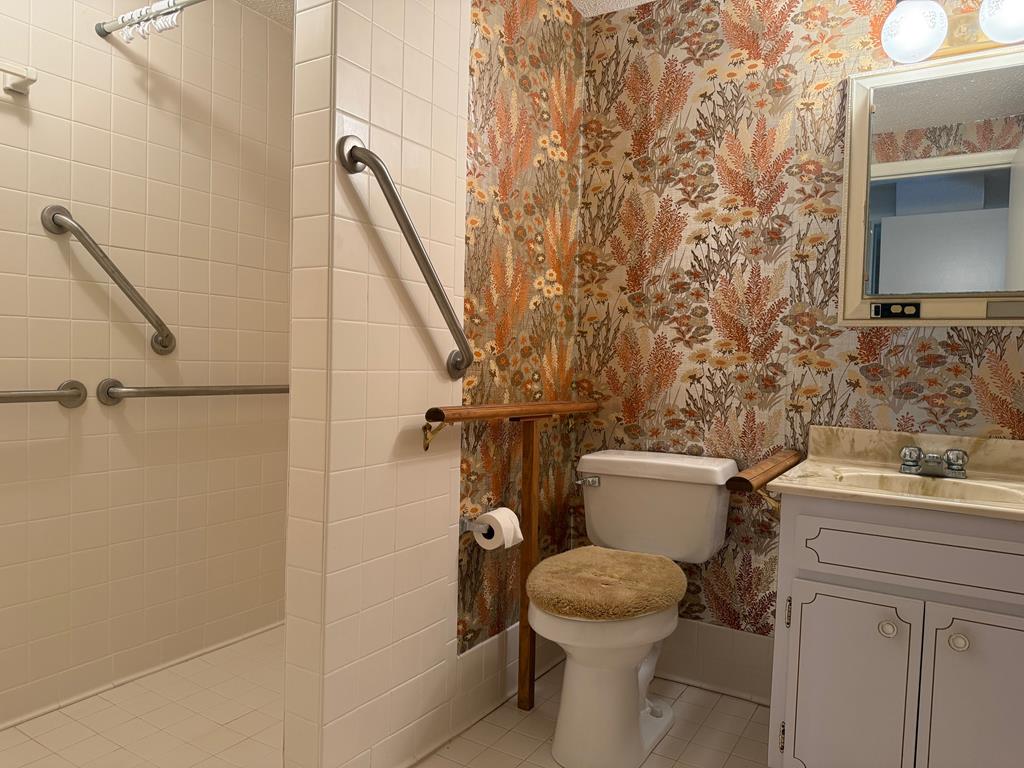 Bathroom with a tiled accessible shower, grab bars, floral wallpaper, toilet, and vanity.