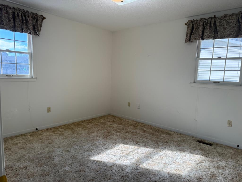 Empty bedroom with beige carpet, two curtained windows, and sunlight across the floor.