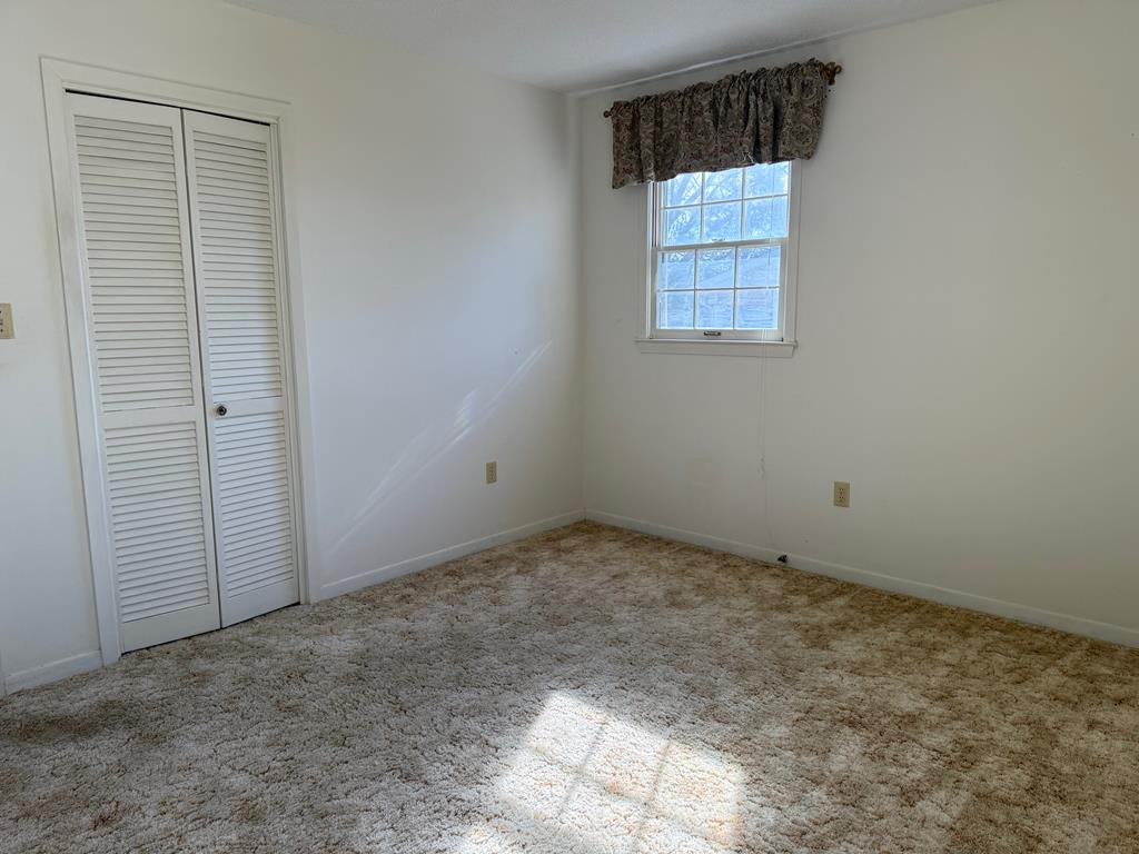 Empty bedroom with beige carpet, louvered closet doors, and a sunlit window.