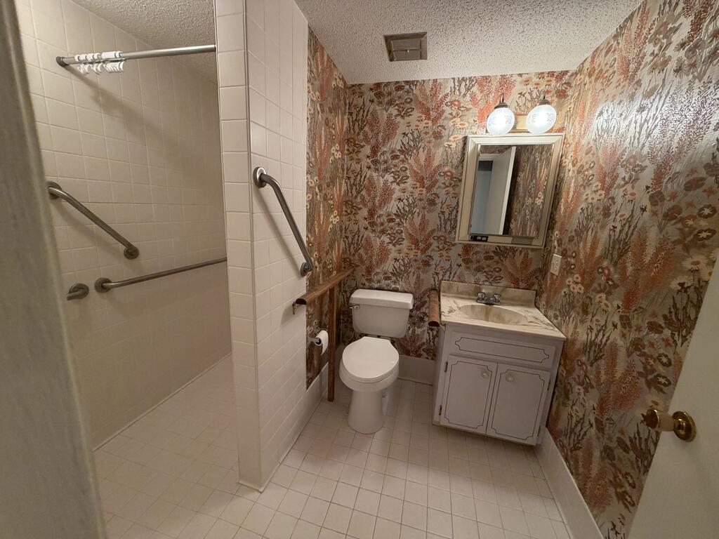 Bathroom with a walk-in tiled shower, grab bars, white vanity, and floral wallpaper.